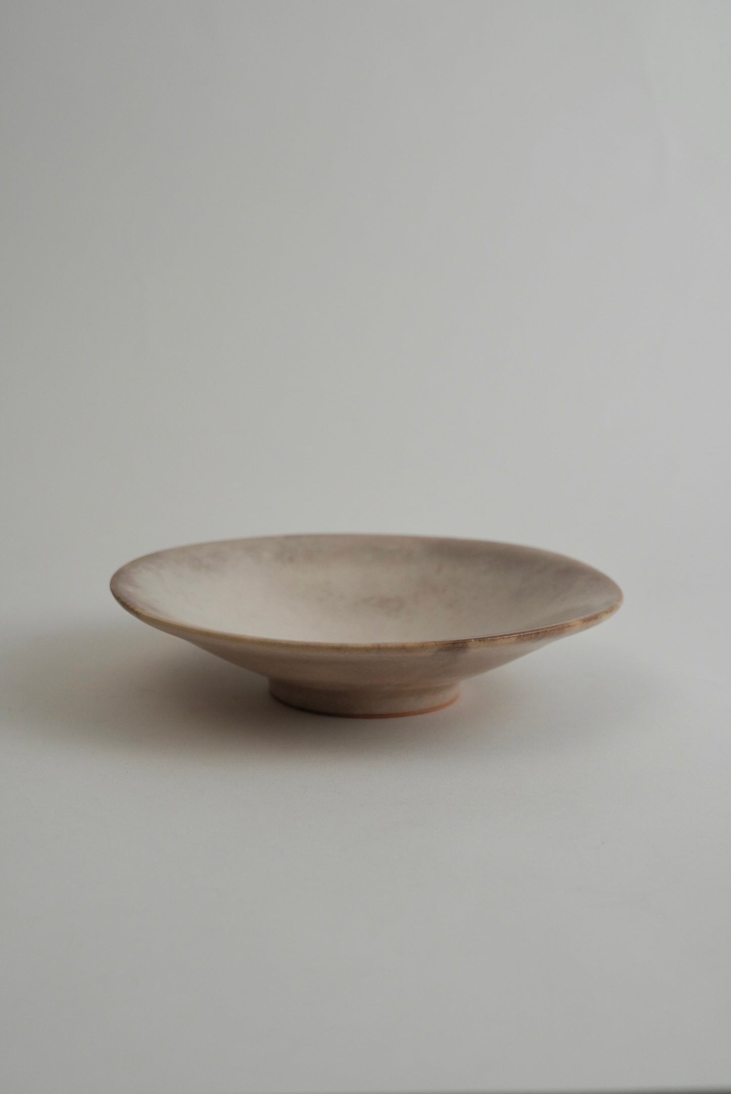 A shallow, wide ceramic dish with a rustic, matte finish in a light beige or off-white glaze, set against a plain light gray background.
