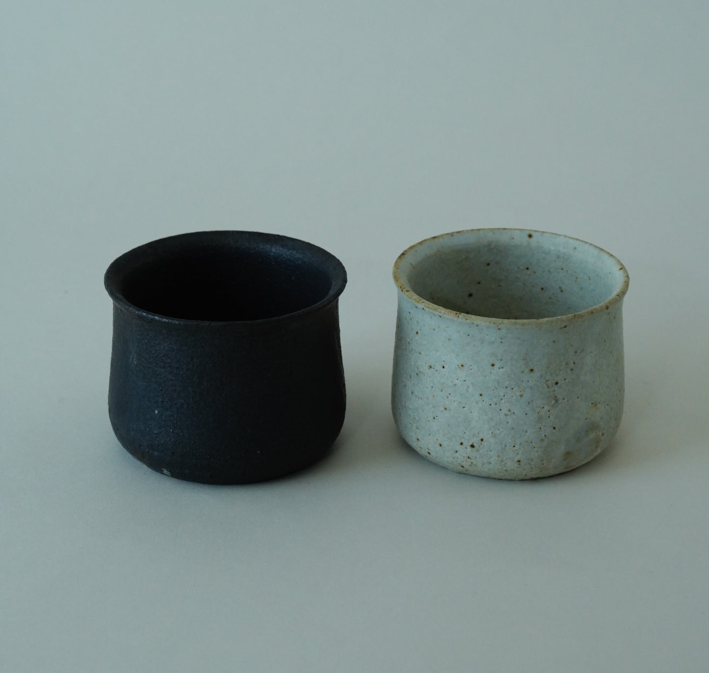 Two ceramic cups, one black and one with a speckled pattern, on a light gray background. Handmade ceramic cup by Japanese artist at Gallery tete Hong Kong.
