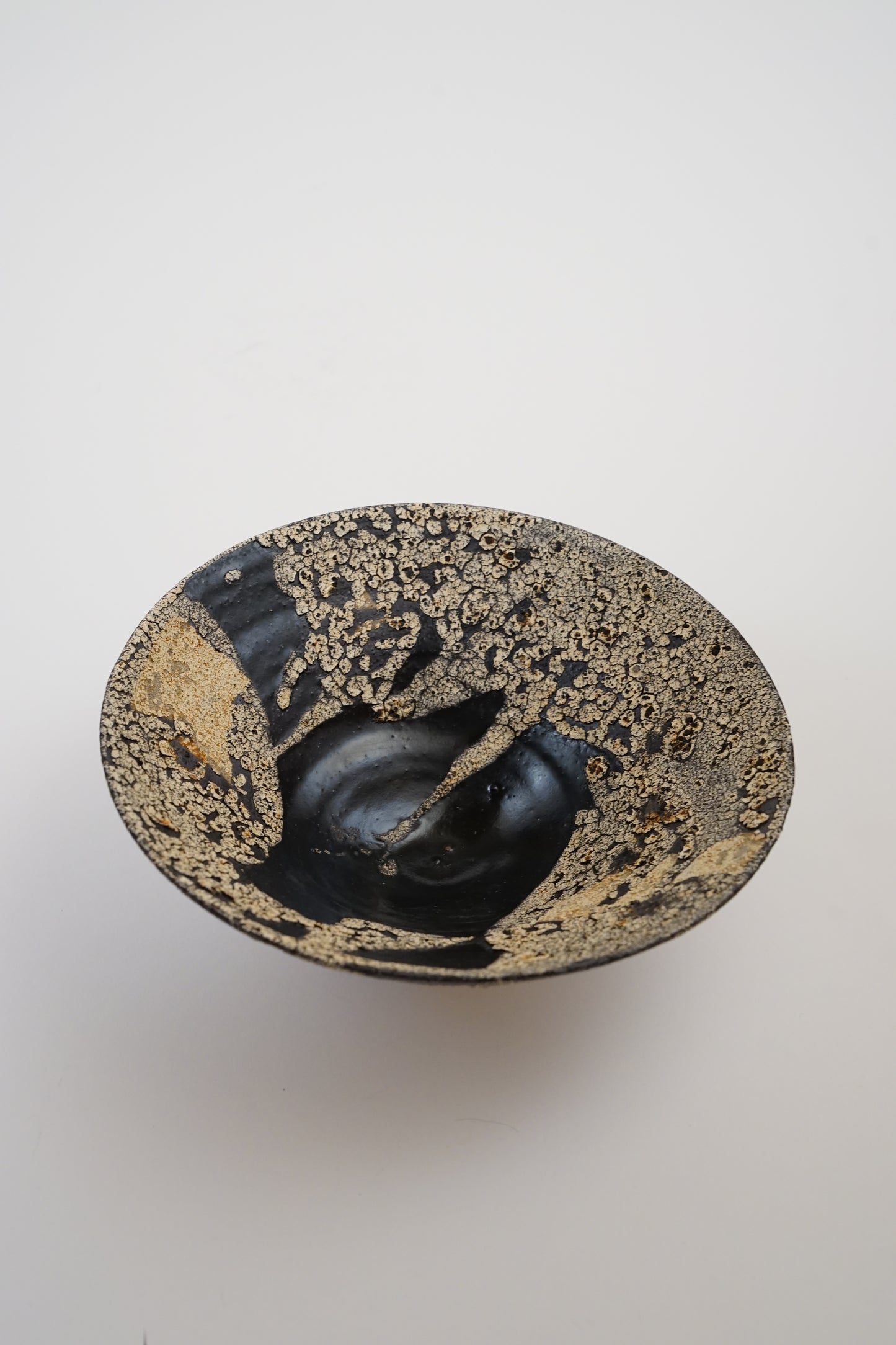 An abstract ceramic bowl with a glossy black center and a textured, metallic gold and bronze speckled rim, photographed from a top-down angle on a light gray background.