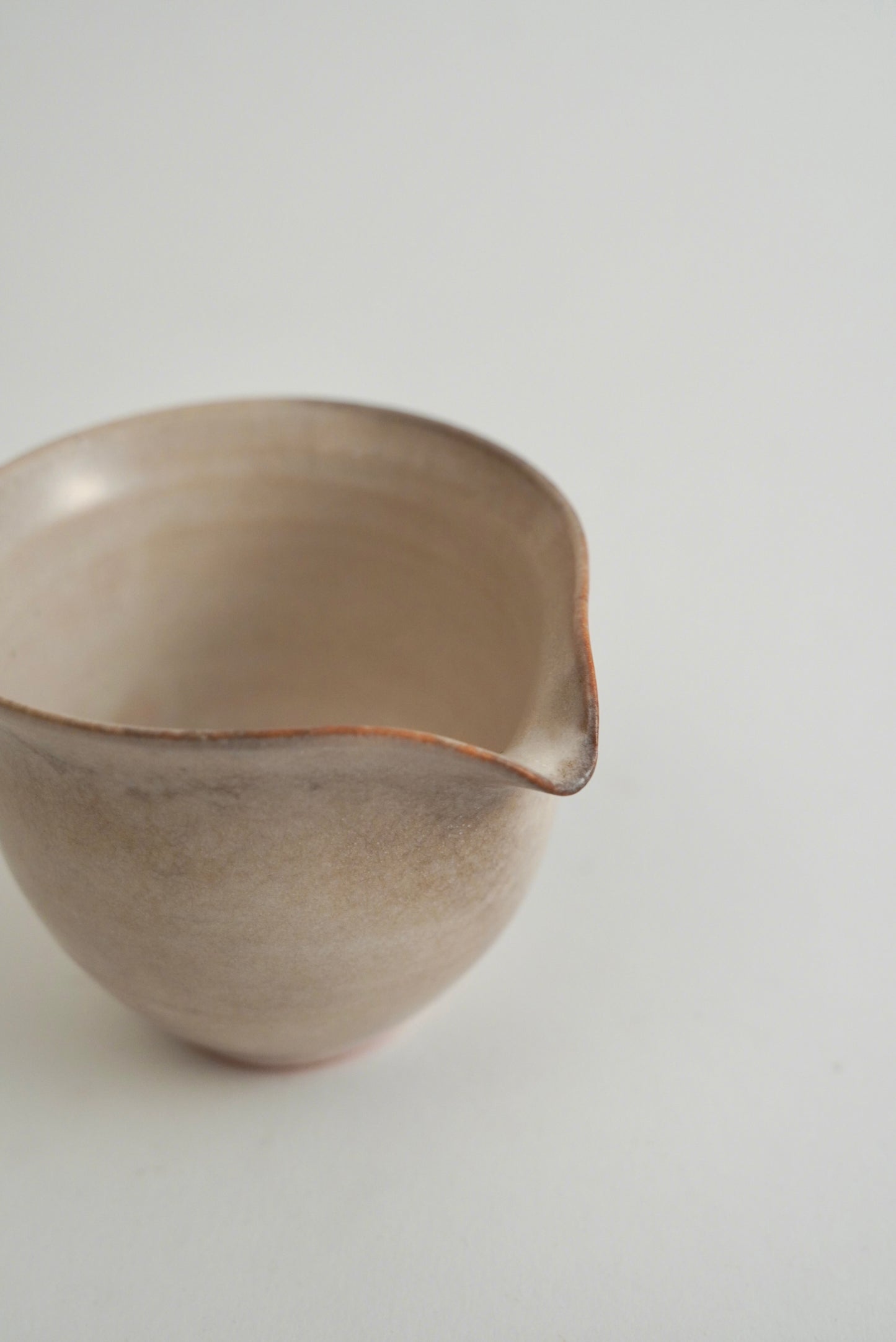 A handmade ceramic pitcher with a rounded body and a small, irregularly shaped spout, featuring a textured matte finish in warm beige and brown tones.