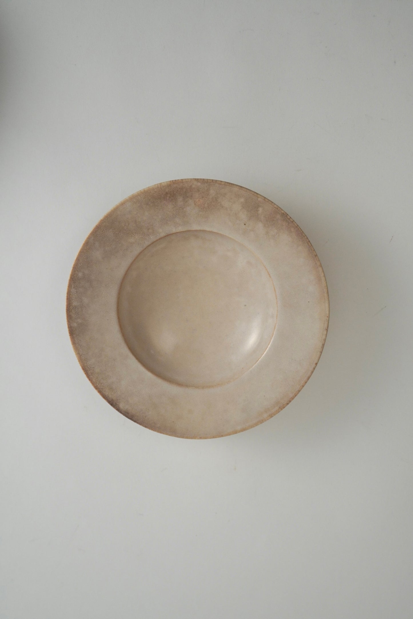 A round, shallow ceramic plate or dish with a rustic, hand-painted finish in a light beige and brown glaze, shown from a top-down perspective on a neutral light gray background.