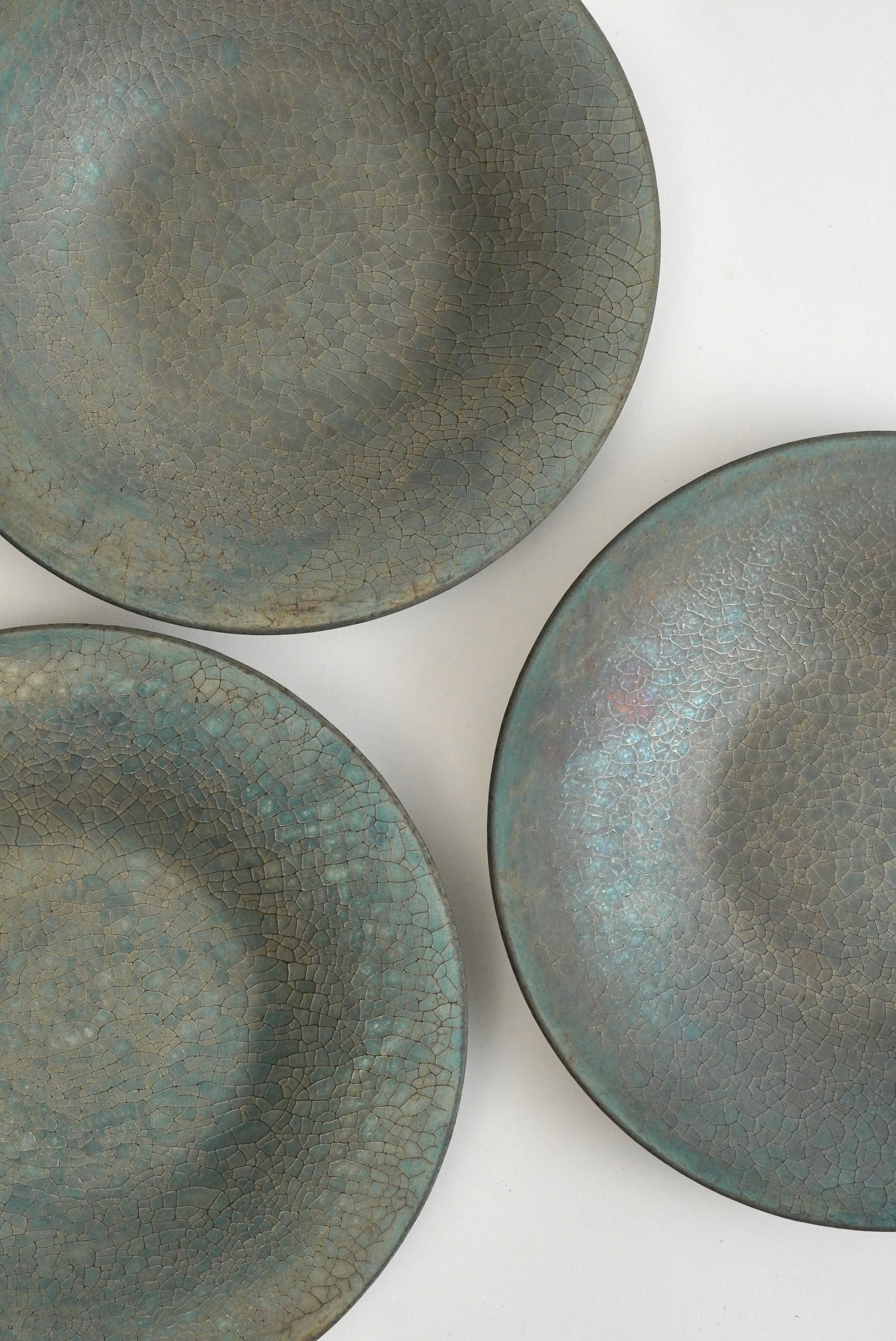 Three ceramic shallow bowls with a textured, crackled glaze in muted blue-green and gray tones, arranged on a plain white background.