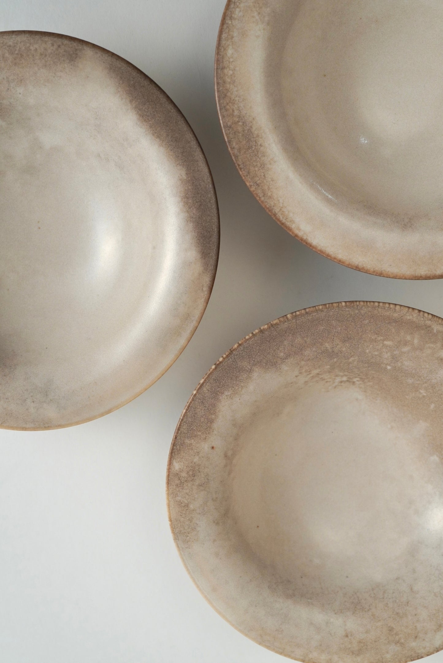 Three ceramic bowls with a rustic, textured matte finish in warm beige and gray tones, arranged diagonally on a light gray background.