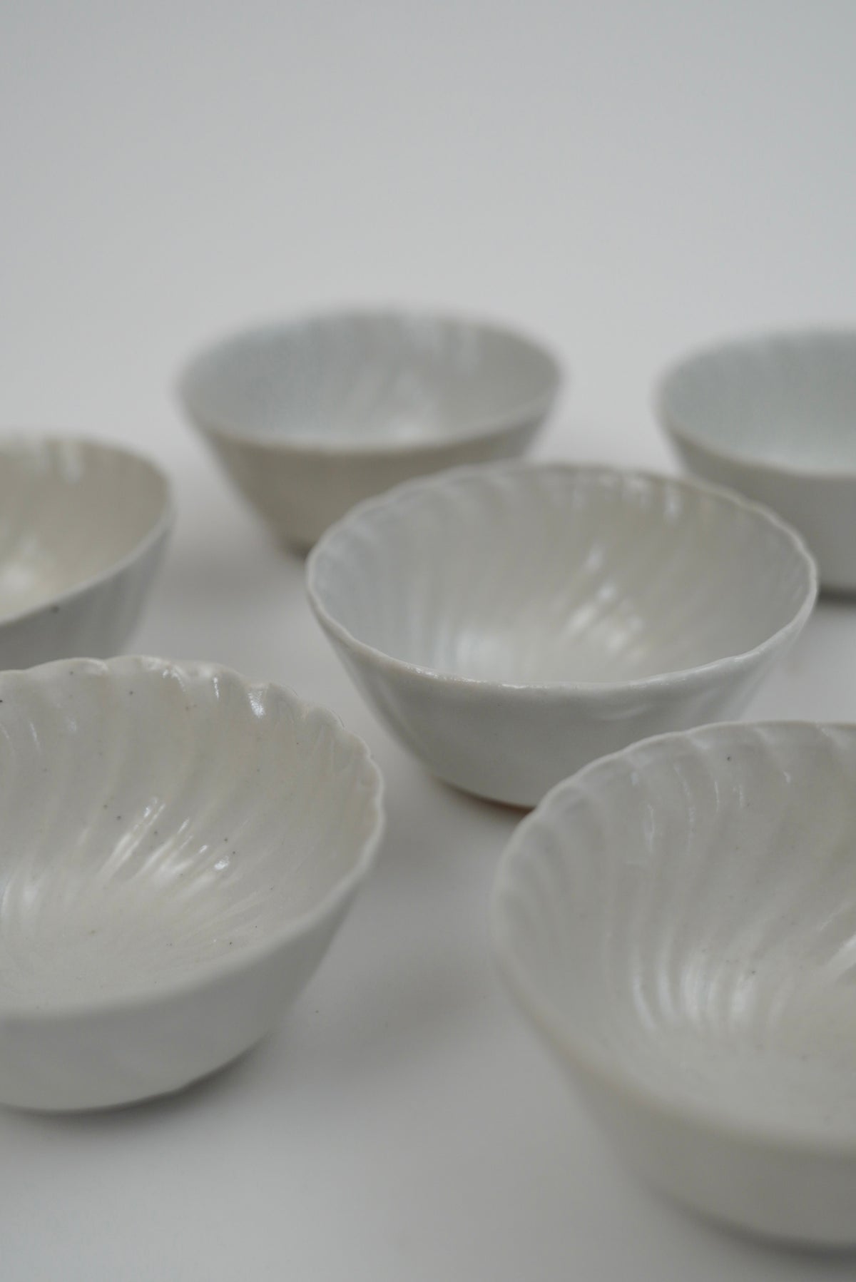 Six small white ceramic bowls with a ribbed, scalloped rim, arranged on a plain light gray background.