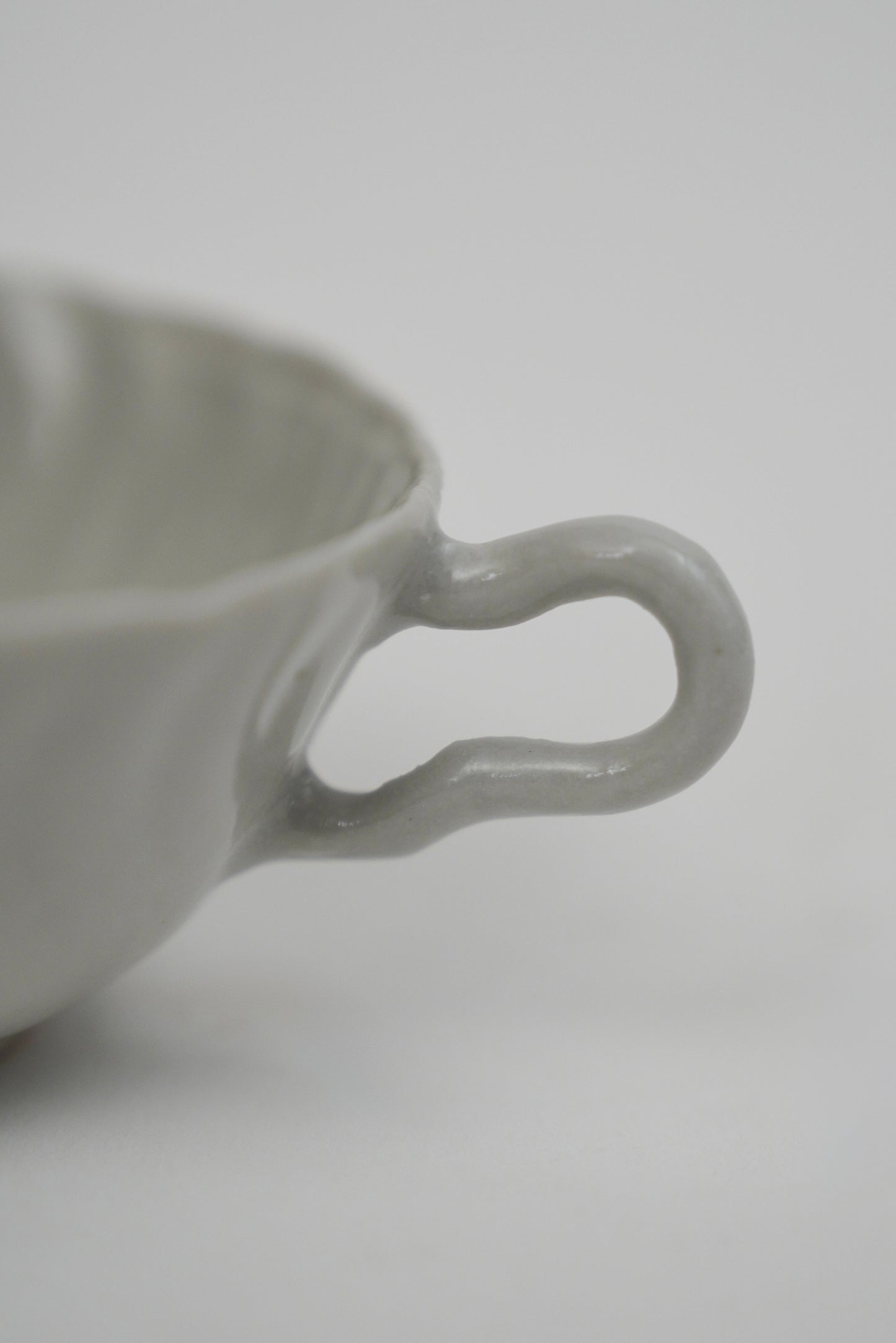 Close-up of a light gray ceramic cup with a wavy, looped handle on a plain light gray background.