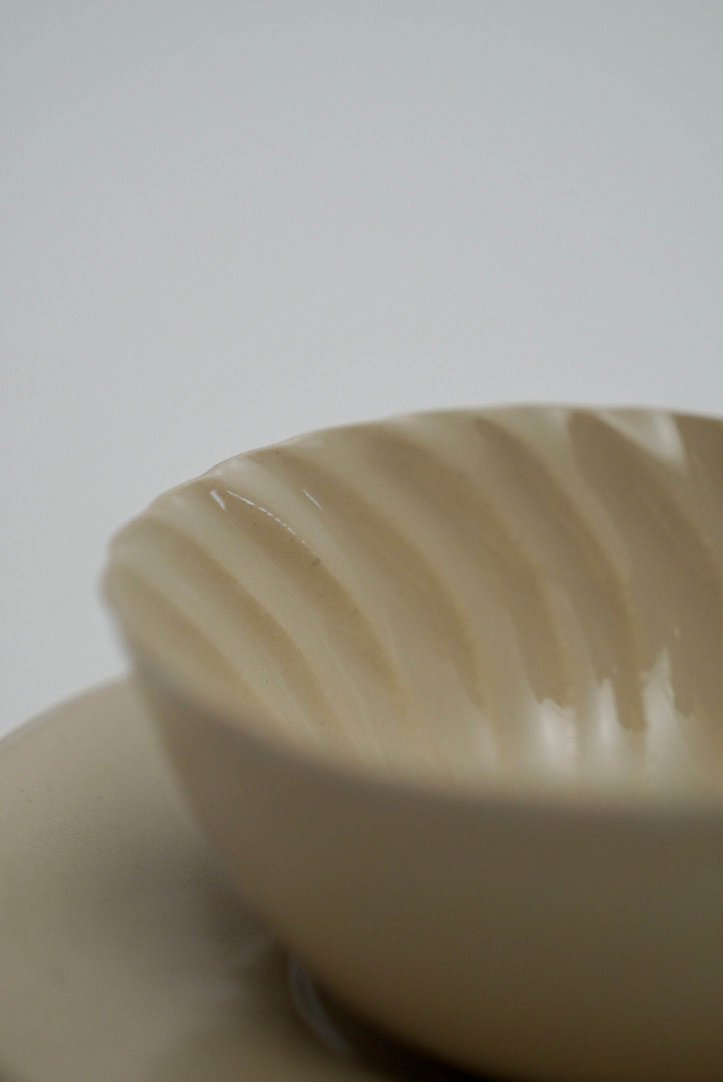 A close-up of a cream-colored ceramic cup with a ribbed, fluted interior, resting on a matching saucer against a neutral light gray background.