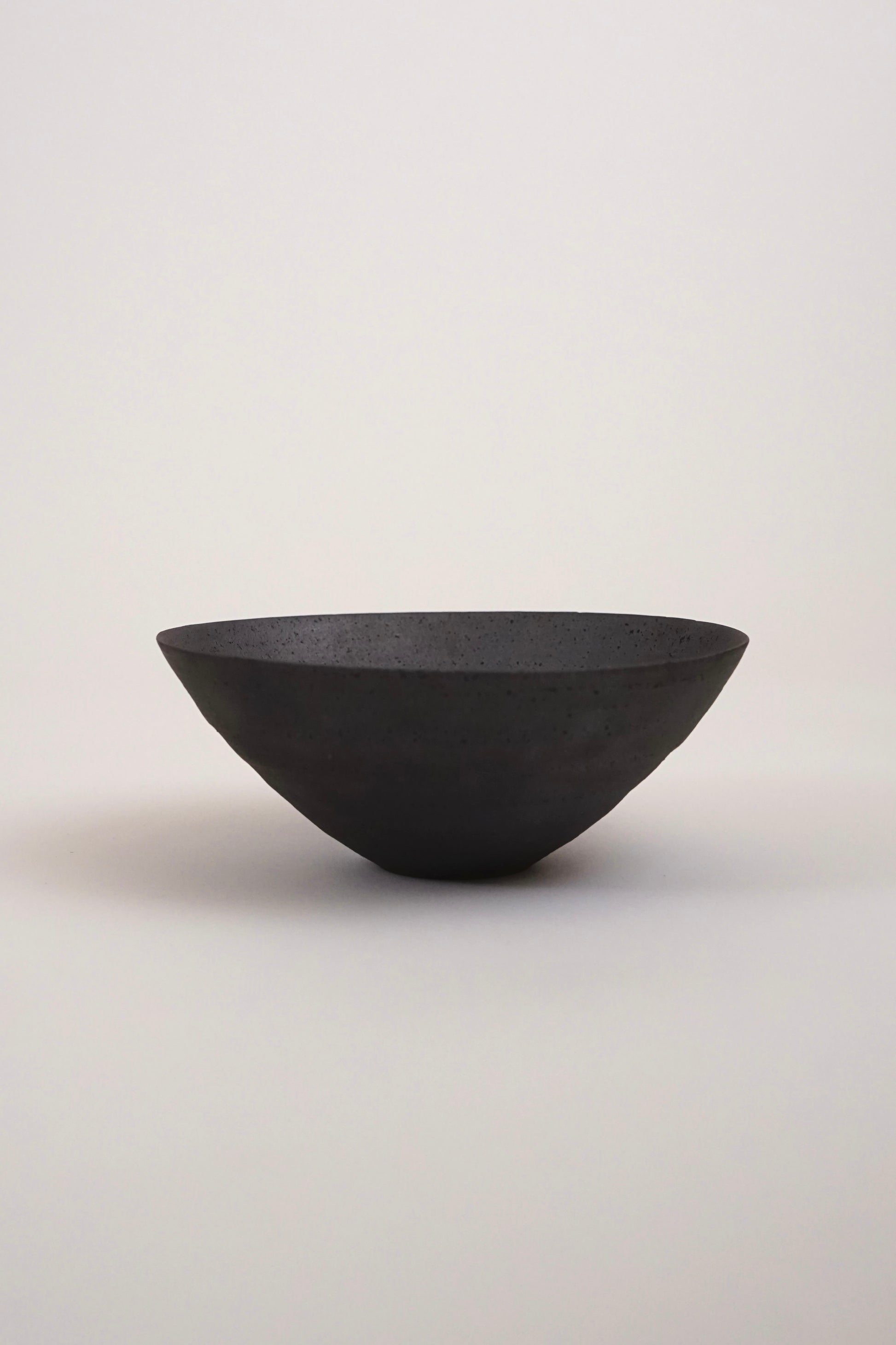 A matte black ceramic bowl with a wide, shallow, wide-brimmed shape, photographed against a neutral light gray background.