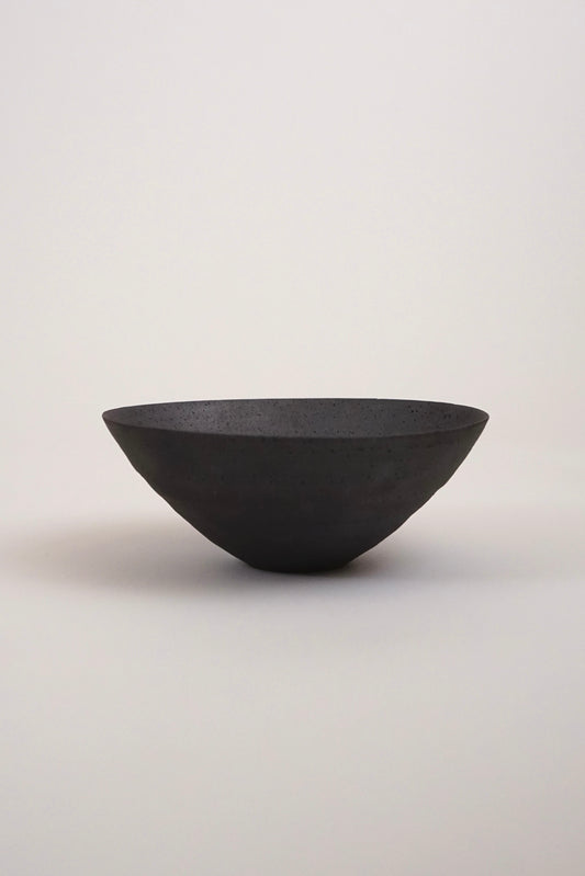 A matte black ceramic bowl with a wide, shallow, wide-brimmed shape, photographed against a neutral light gray background.