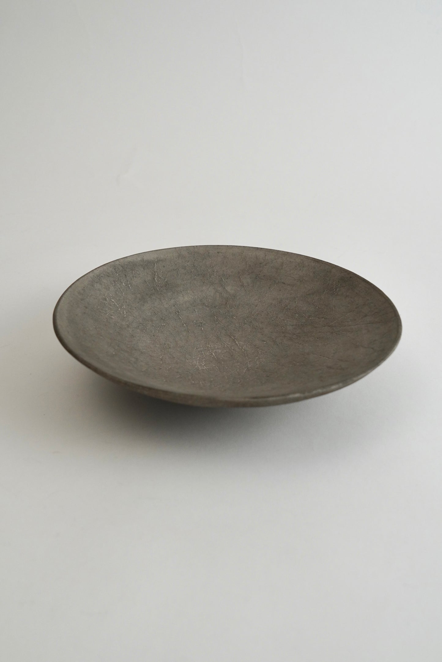 A shallow, round ceramic plate with a textured matte finish in a dark gray color, photographed against a neutral light gray background.