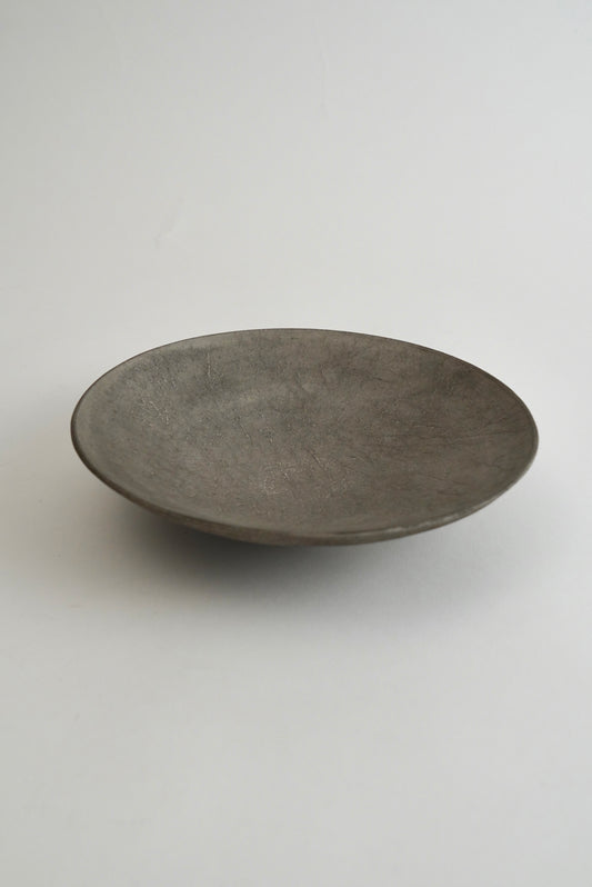 A shallow, round ceramic plate with a textured matte finish in a dark gray color, photographed against a neutral light gray background.