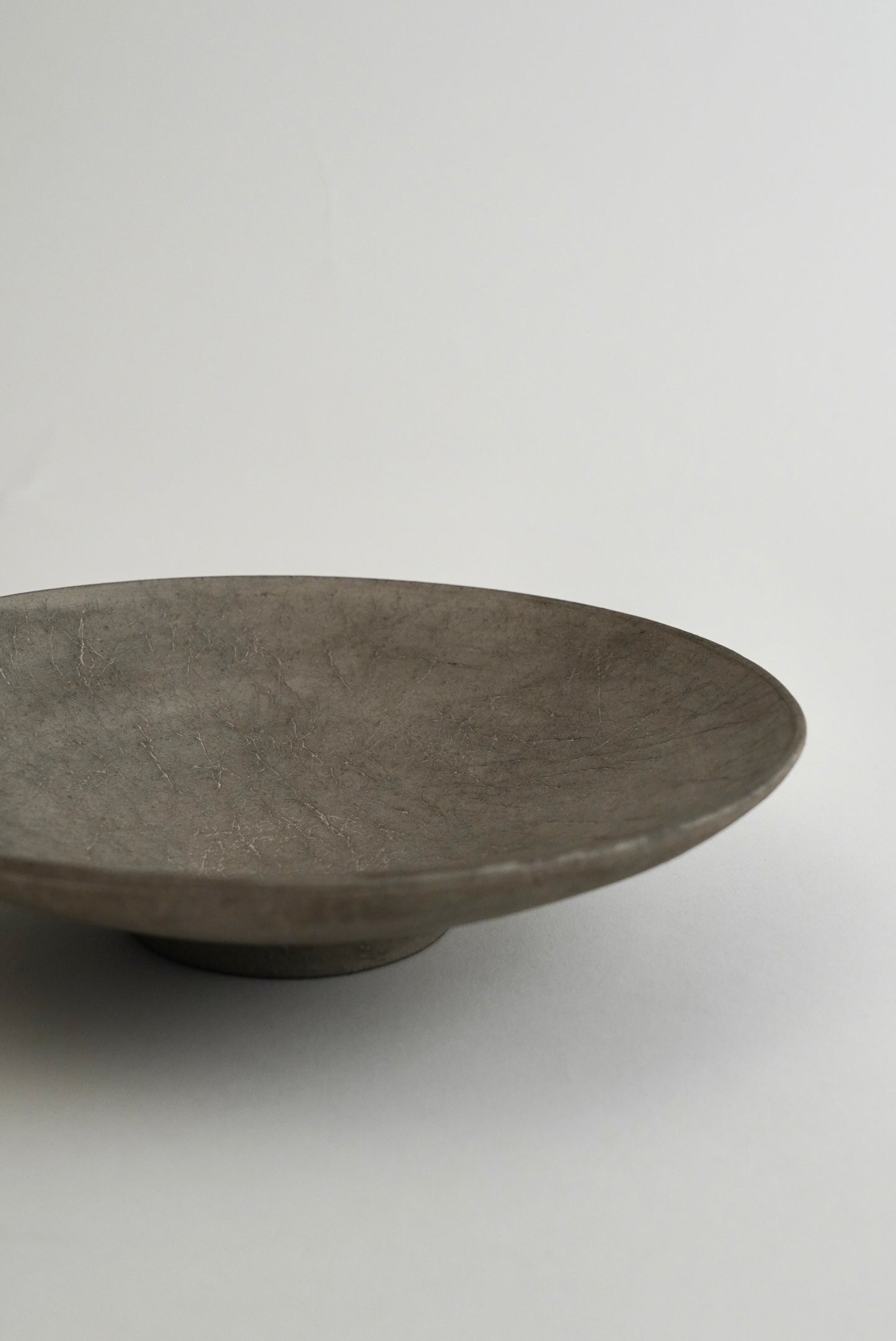 A minimalist, shallow round ceramic plate or bowl in a textured matte gray finish, photographed on a seamless light gray background.