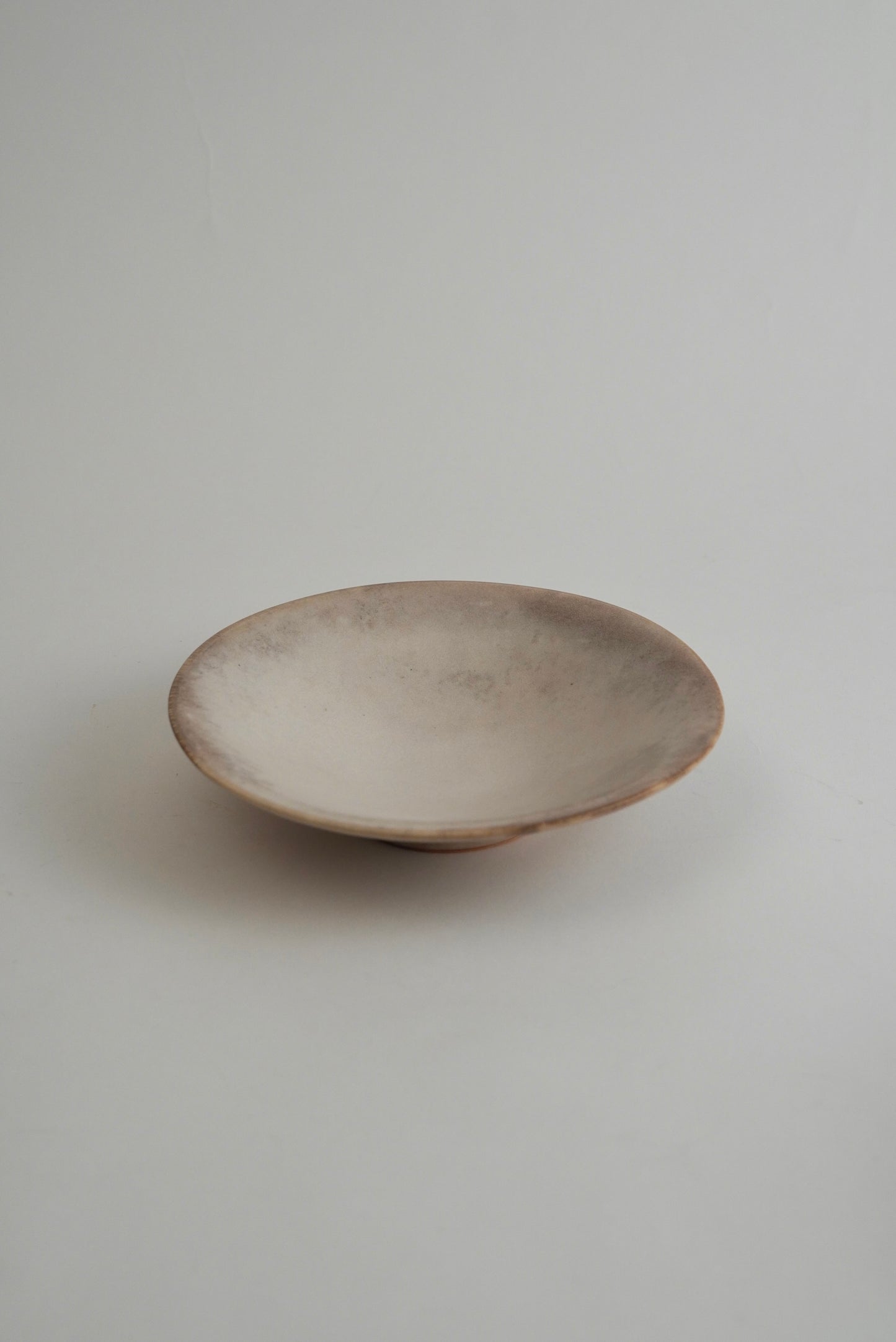 A shallow, wide ceramic plate or dish with a rustic, hand-painted finish in earthy beige and brown tones, set against a plain light gray background.