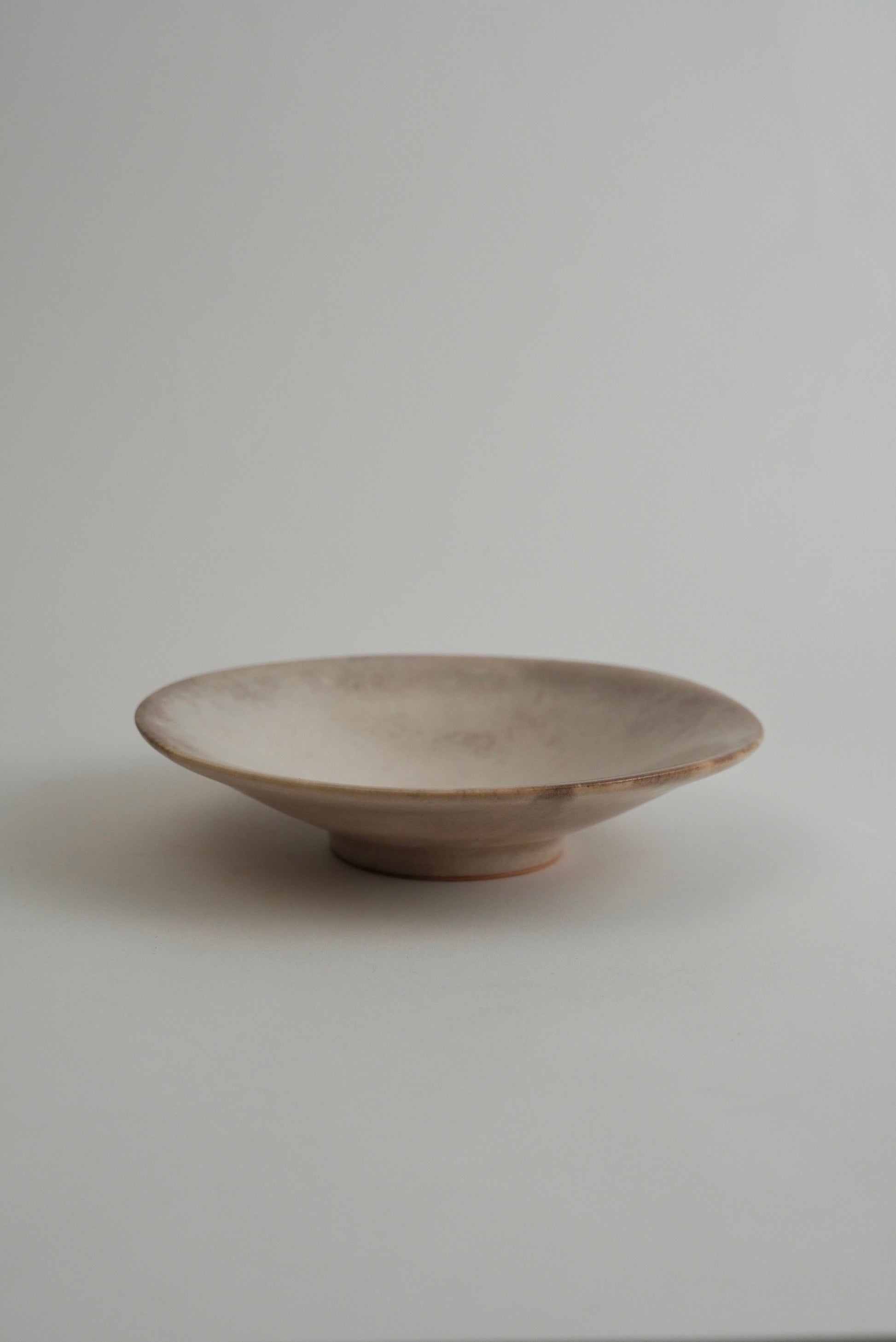 A shallow, wide ceramic dish with a rustic, matte finish in a light beige or off-white glaze, set against a plain light gray background.