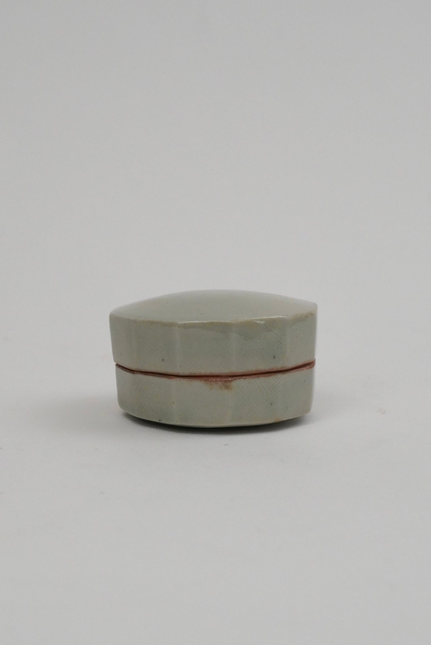 A small hexagonal ceramic box with a pale greenish-gray glaze and a thin reddish-brown band around the middle, photographed on a plain light gray background.