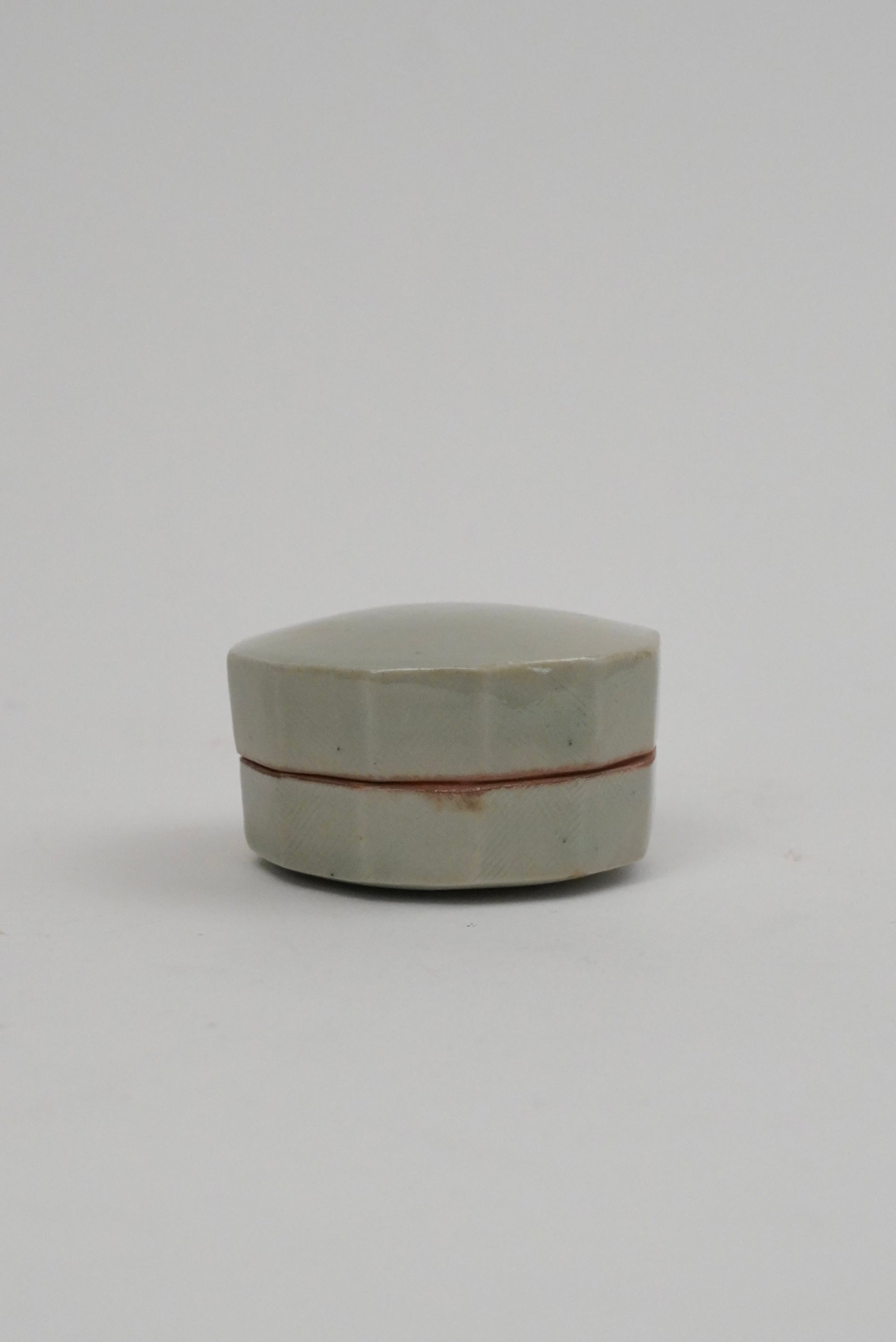 A small hexagonal ceramic box with a pale greenish-gray glaze and a thin reddish-brown band around the middle, photographed on a plain light gray background.