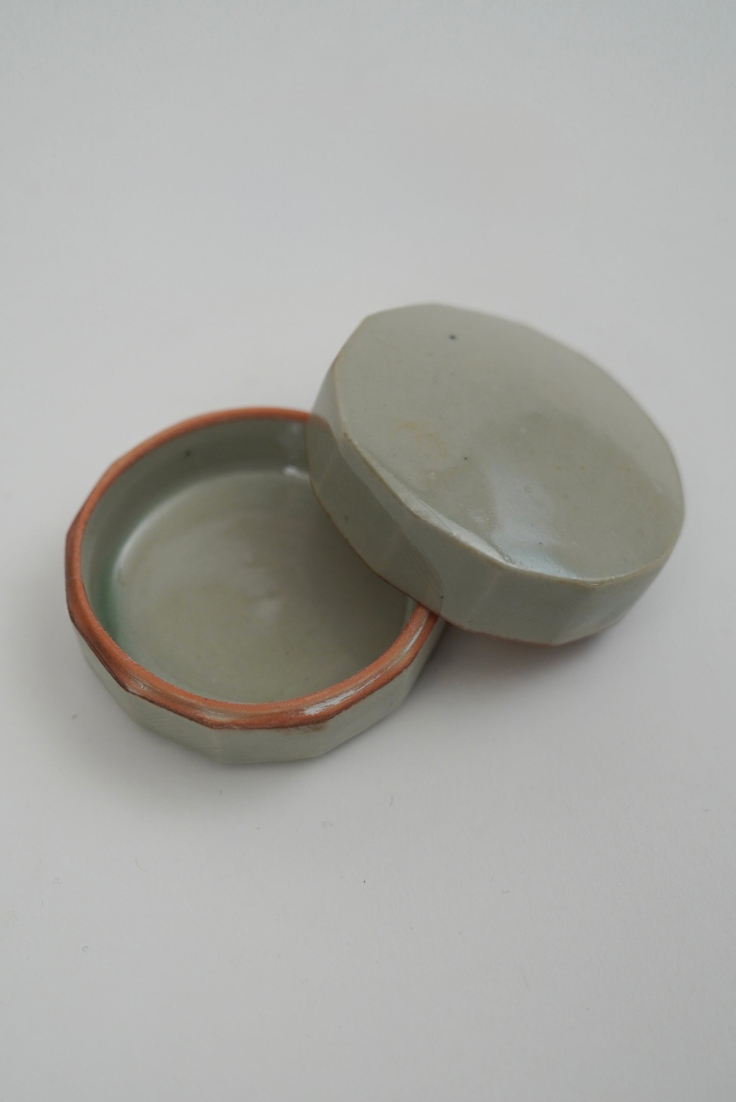 A small handmade ceramic box with a matching lid, featuring a pale sage green glaze and a contrasting terracotta rim, set against a plain light gray background.