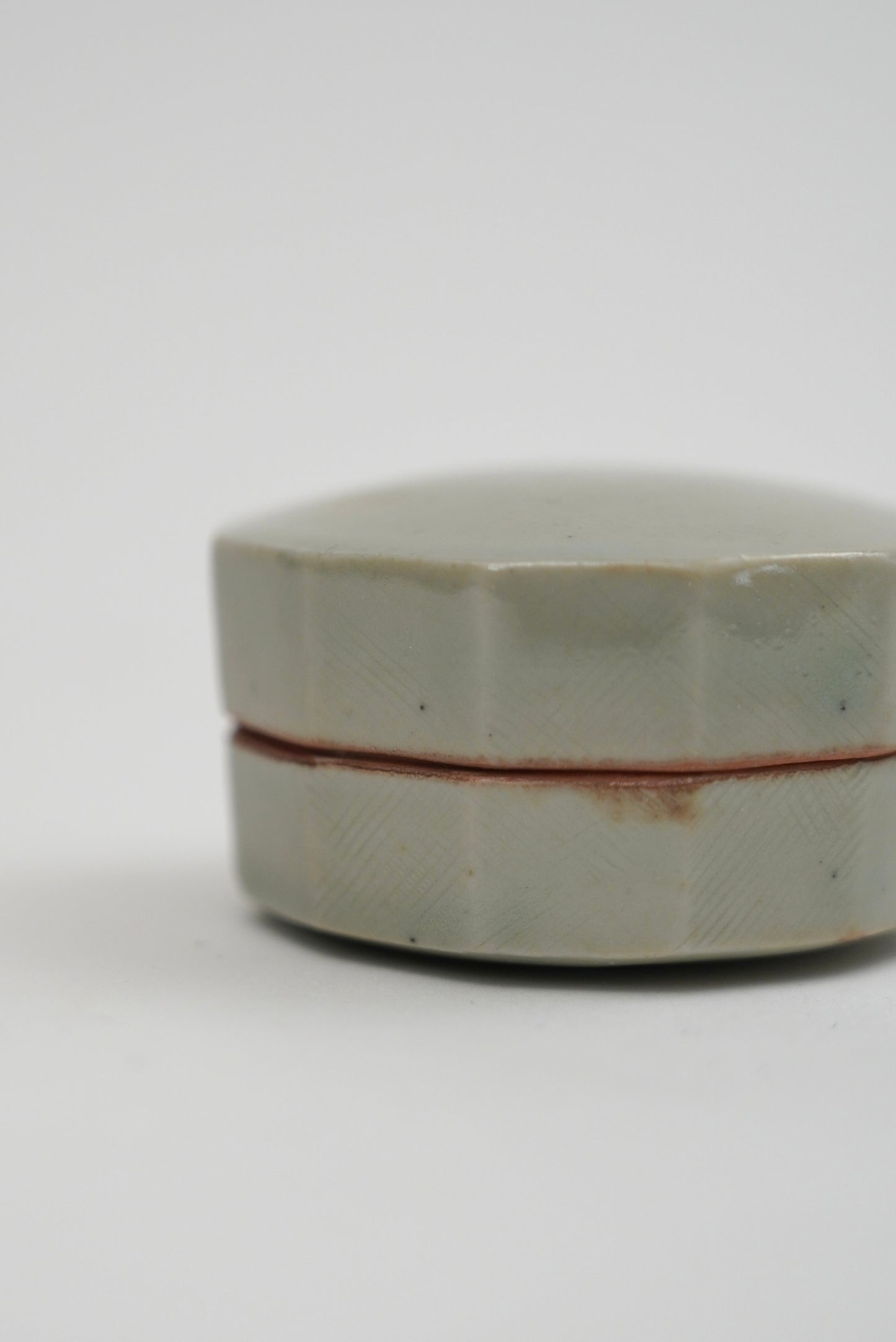 A small round ceramic box with a faceted lid, featuring a pale celadon green glaze and a thin reddish-brown seam line, set against a plain light gray background.