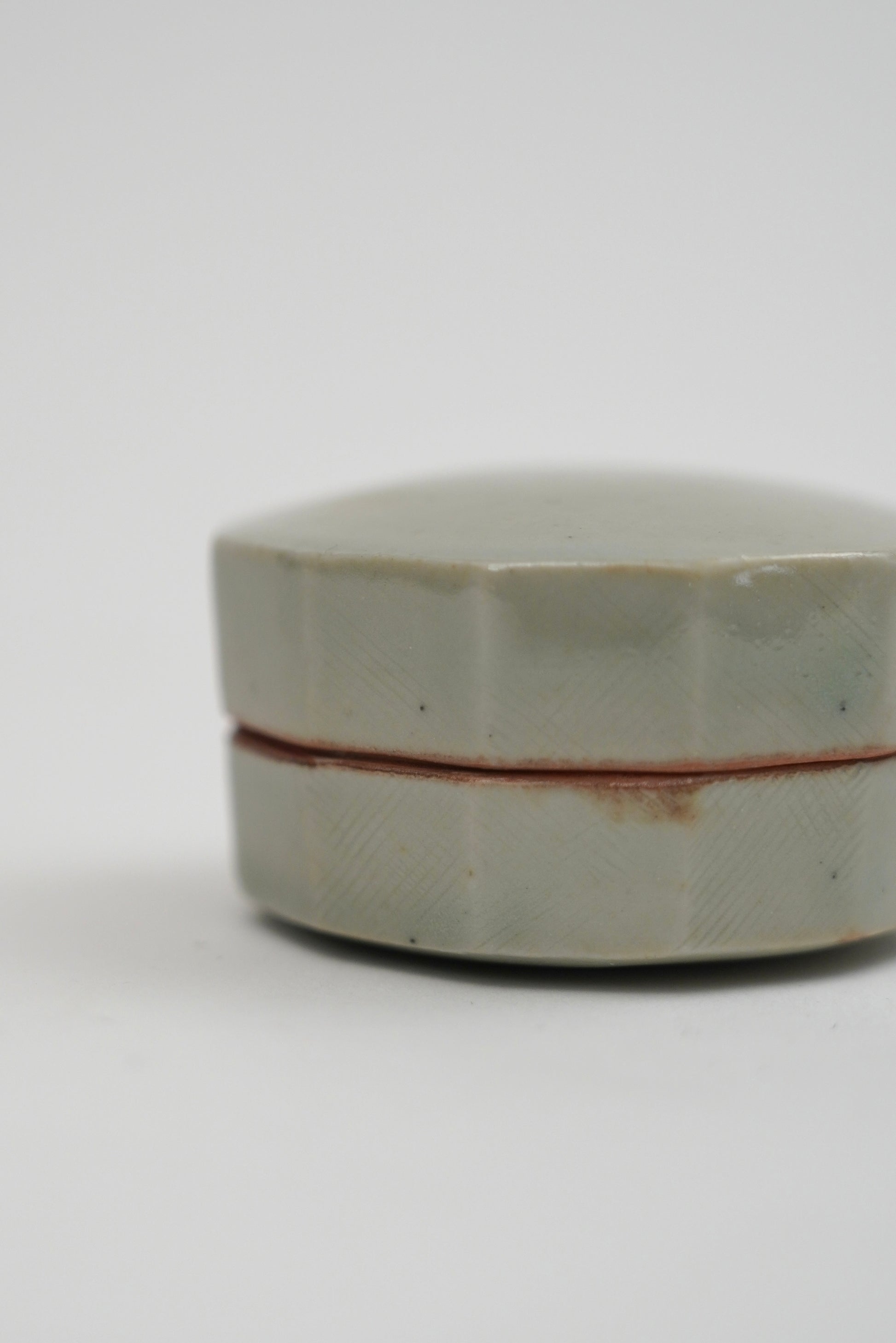 A small round ceramic box with a faceted lid, featuring a pale celadon green glaze and a thin reddish-brown seam line, set against a plain light gray background.