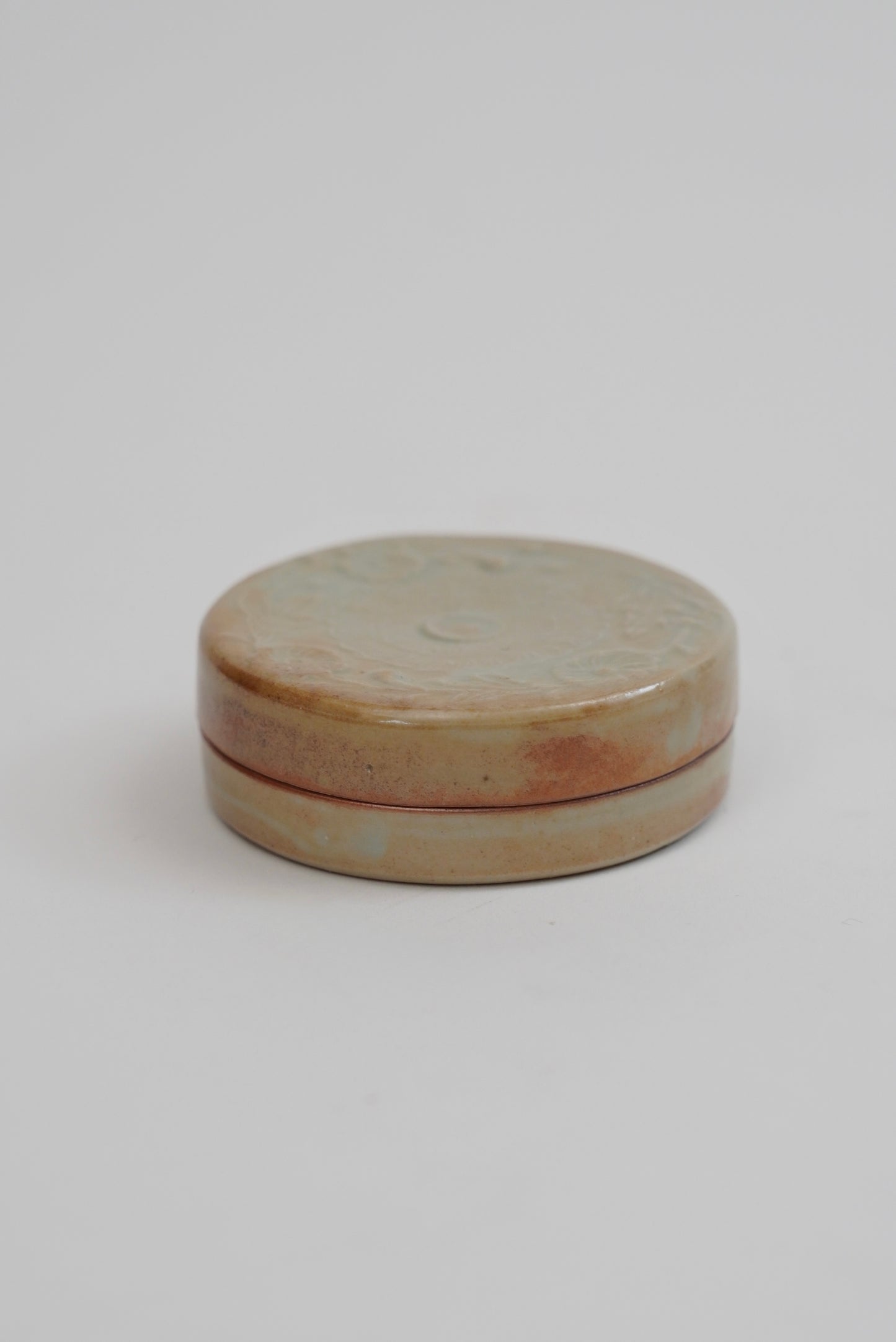 A small round ceramic box with a lid, featuring a glazed finish in earthy beige, brown, and soft pink tones, set against a plain light gray background.