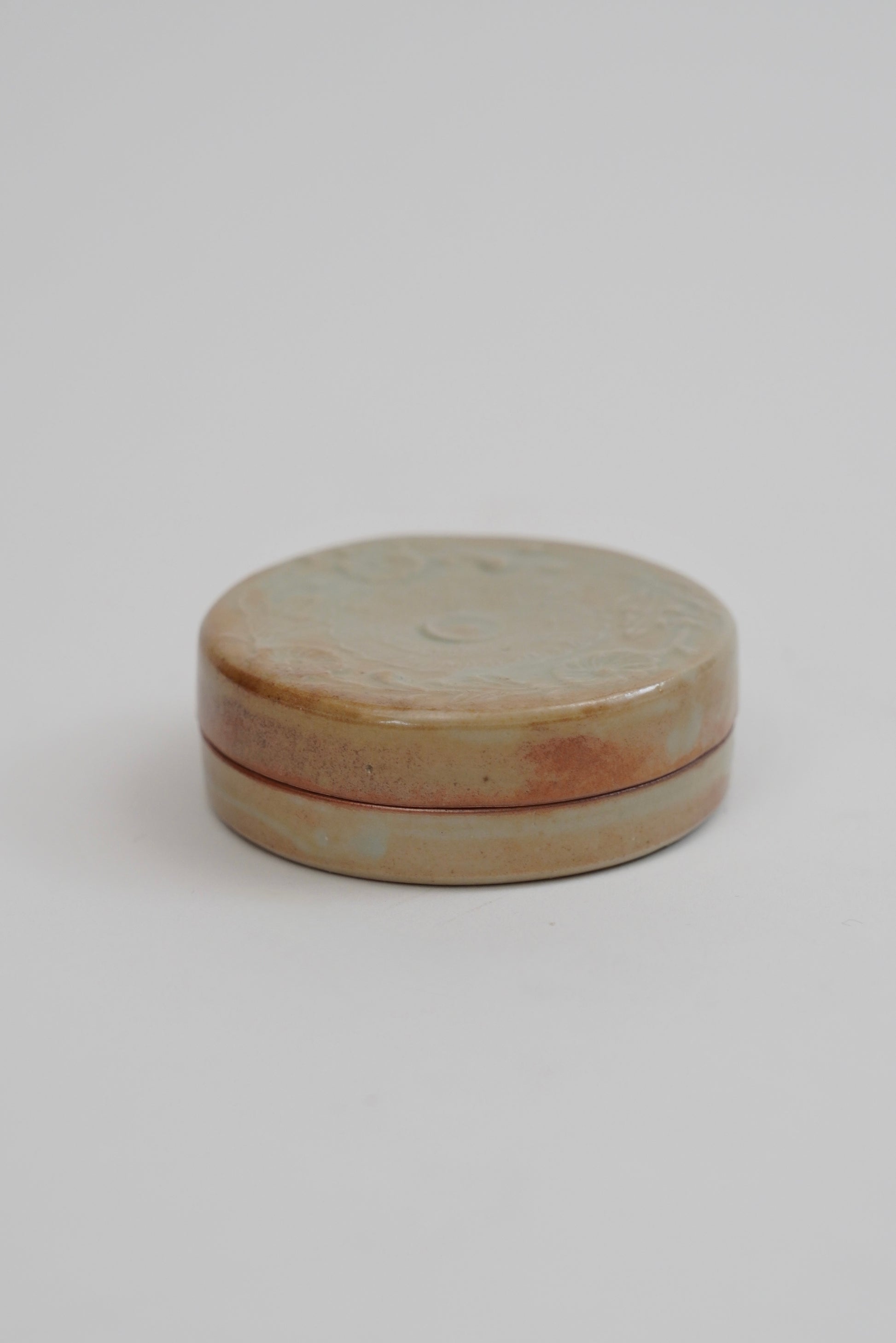 A small round ceramic box with a lid, featuring a glazed finish in earthy beige, brown, and soft pink tones, set against a plain light gray background.