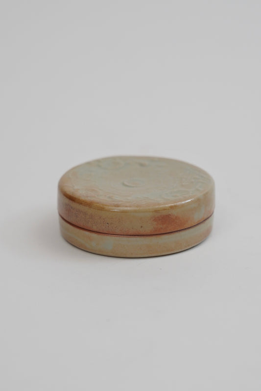 A small round ceramic box with a lid, featuring a glazed finish in earthy beige, brown, and soft pink tones, set against a plain light gray background.