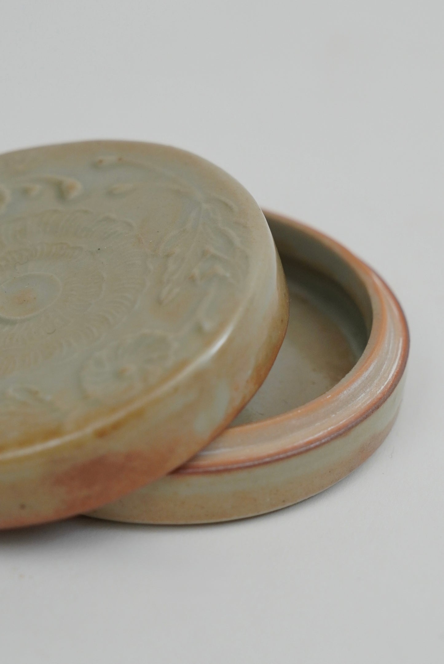 A close-up of a round ceramic lidded container with a slightly raised rim, shown on a plain light background. The exterior glaze is a muted warm gray-beige with soft brown variation, and the interior appears pale pinkish-beige.