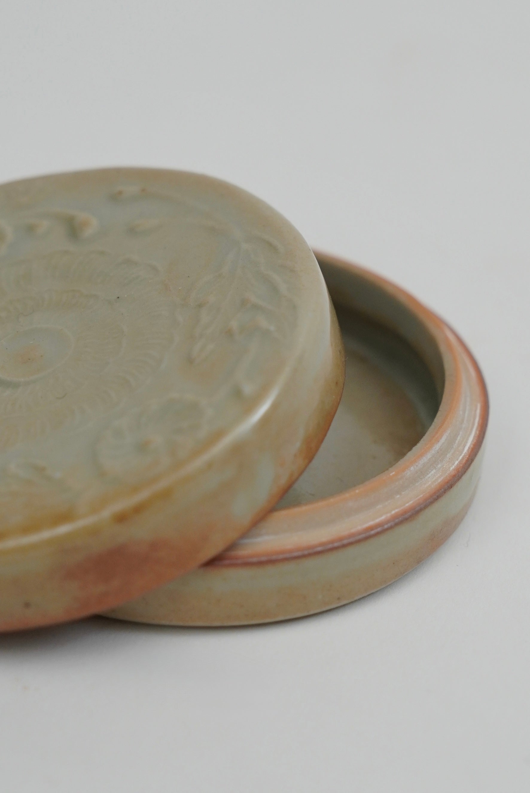 A close-up of a round ceramic lidded container with a slightly raised rim, shown on a plain light background. The exterior glaze is a muted warm gray-beige with soft brown variation, and the interior appears pale pinkish-beige.