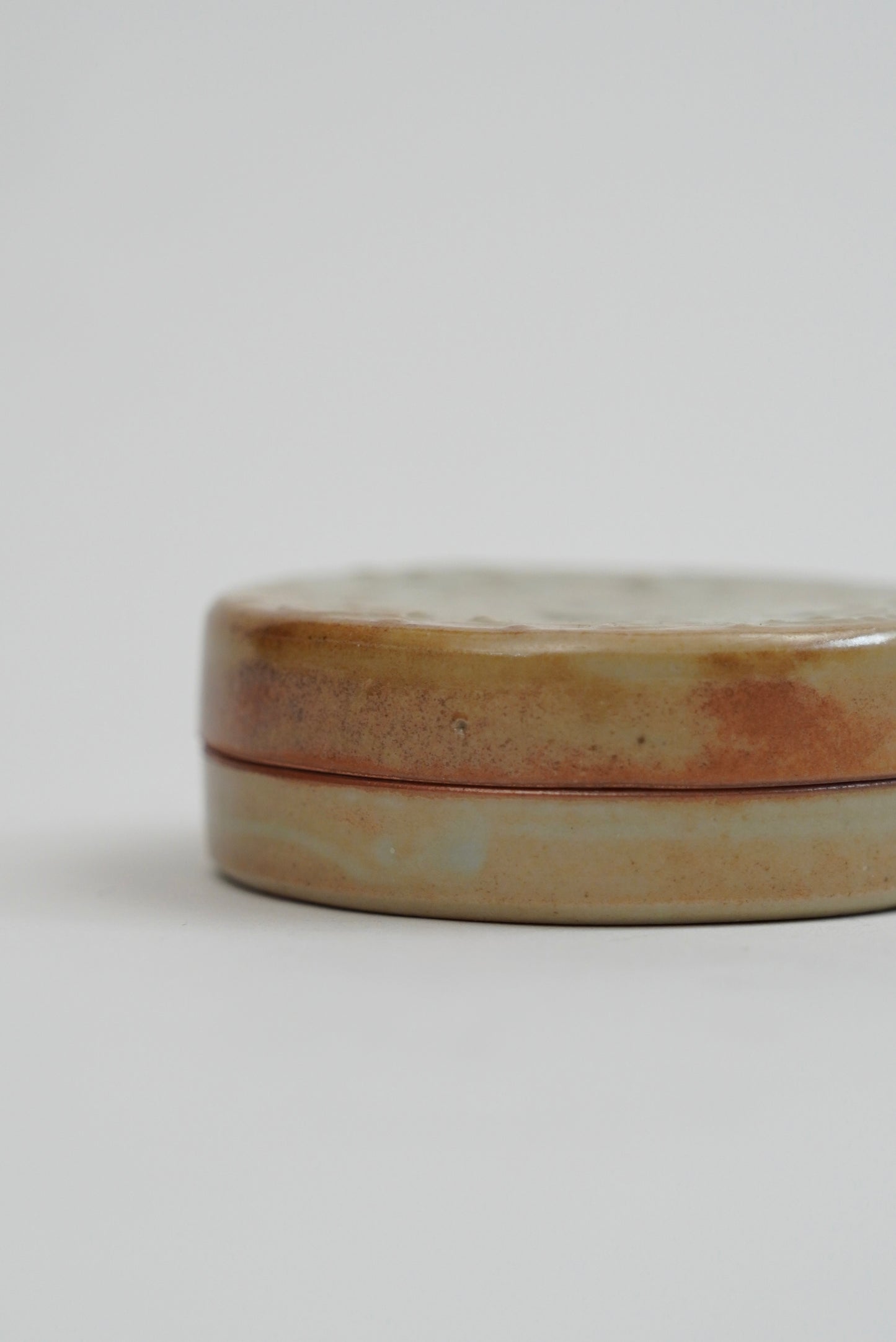 A small round ceramic tin with a lid, featuring a rustic glaze in earthy beige, brown, and soft pink tones, photographed on a clean light gray background.