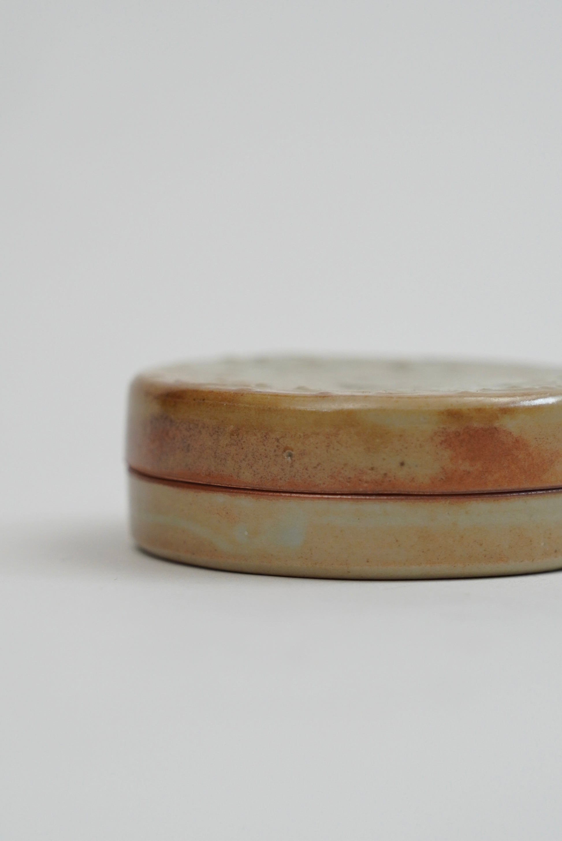 A small round ceramic tin with a lid, featuring a rustic glaze in earthy beige, brown, and soft pink tones, photographed on a clean light gray background.