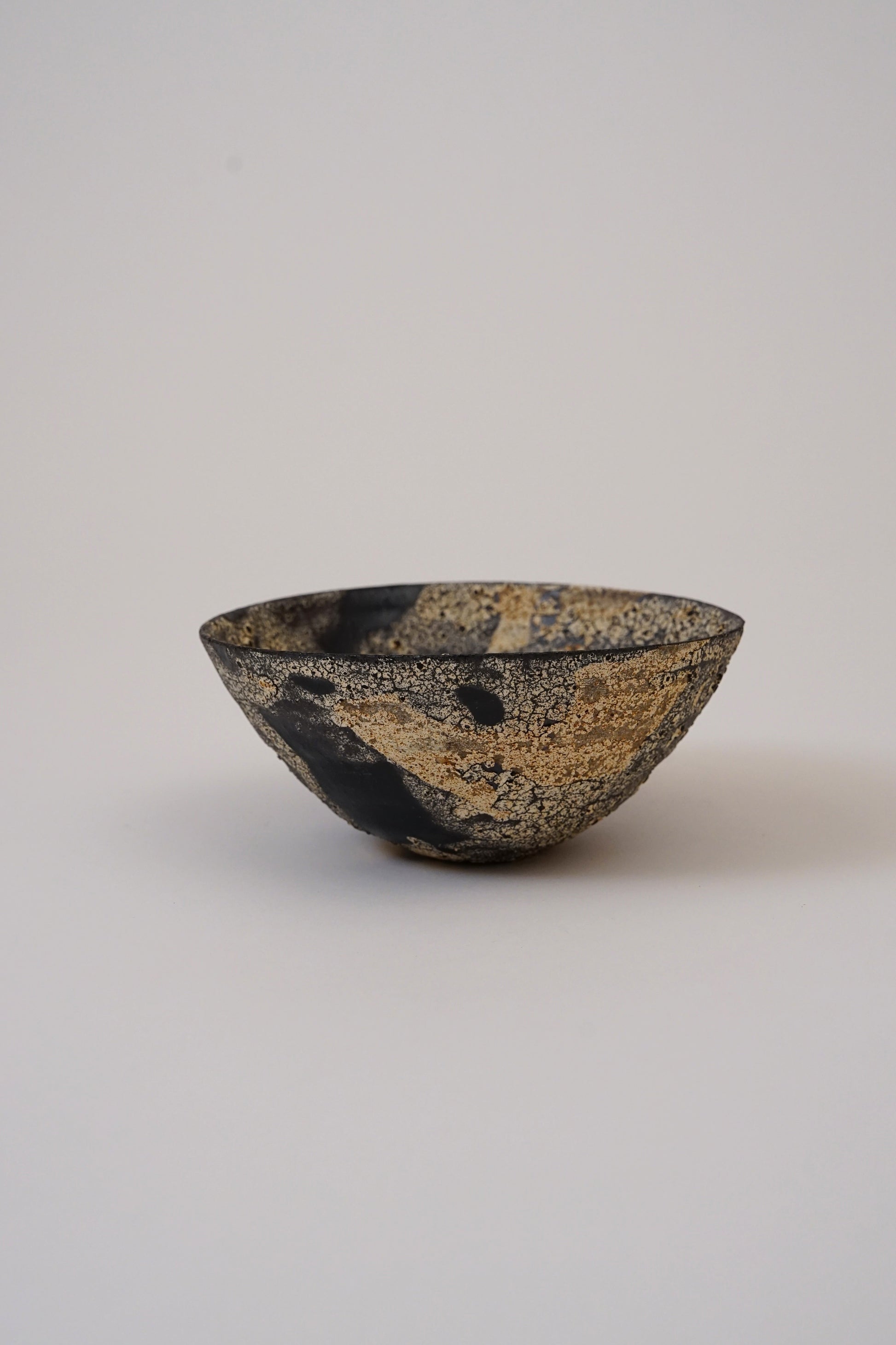 A small handmade ceramic bowl with a textured black and gold speckled glaze, photographed against a neutral light gray background.