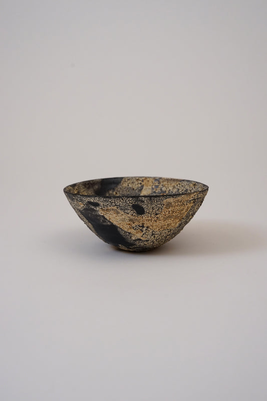 A small handmade ceramic bowl with a textured black and gold speckled glaze, photographed against a neutral light gray background.