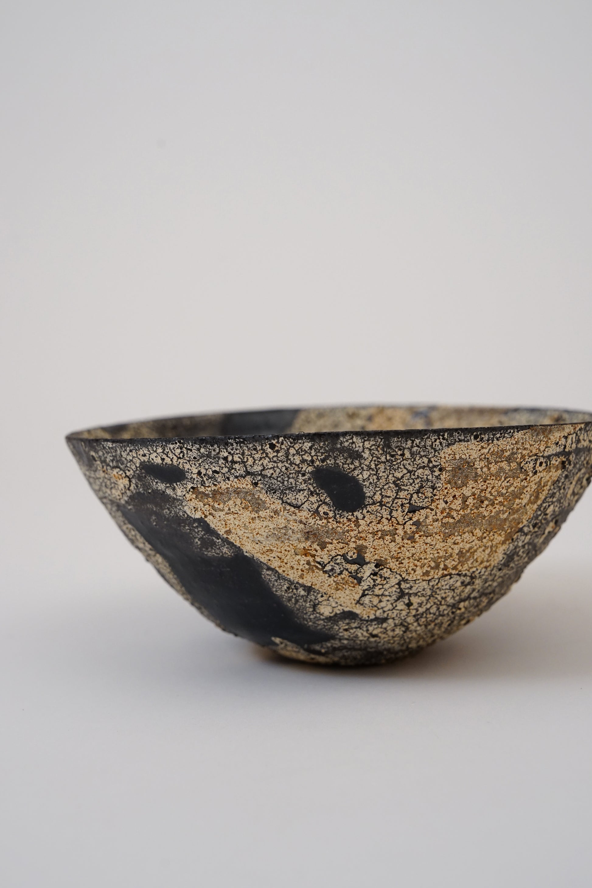 A handmade ceramic bowl with a textured, crackled glaze in black and metallic gold, photographed on a neutral light gray background.