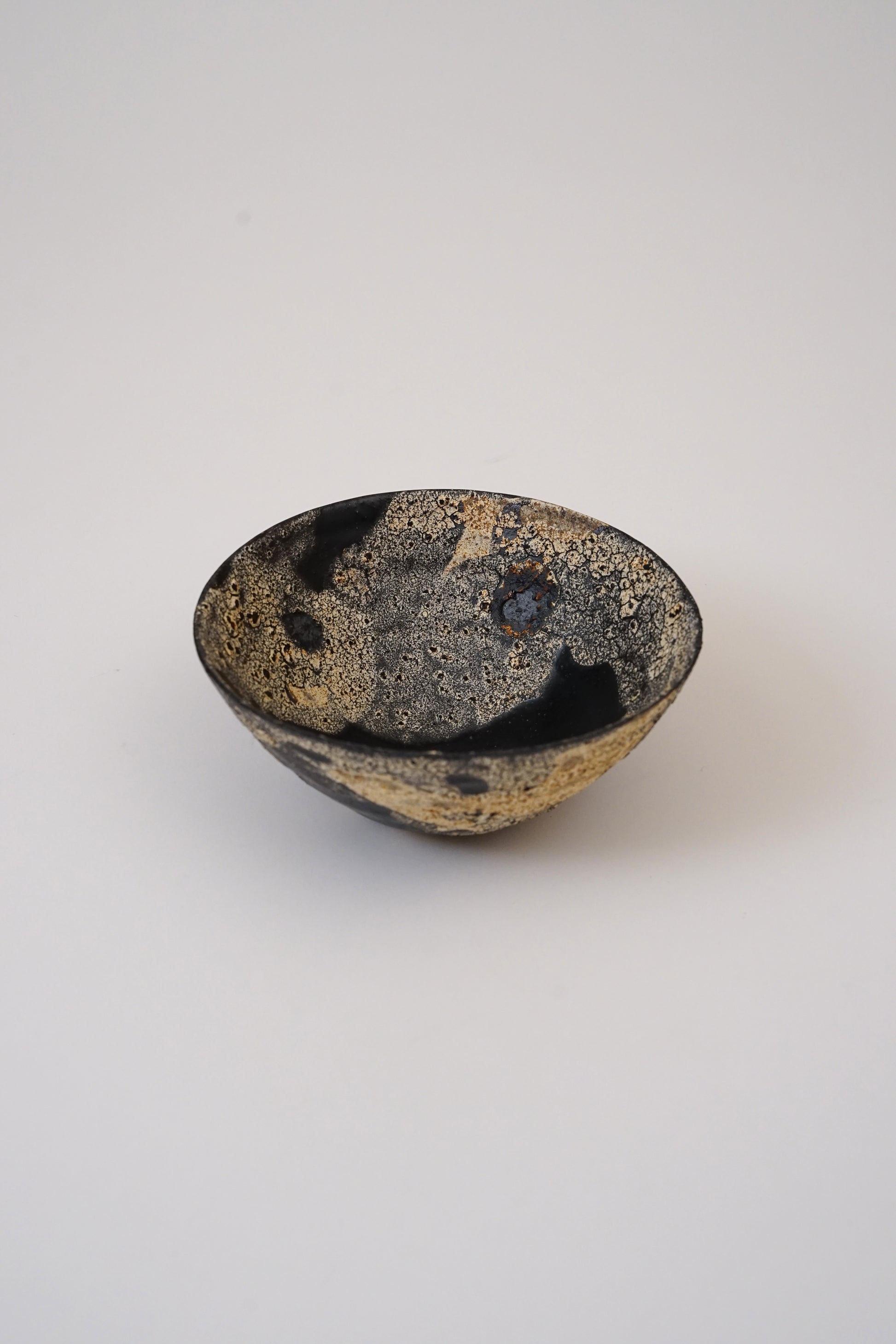 A small handmade ceramic bowl with a textured, speckled glaze in black, gold, and brown tones, photographed on a neutral light gray background.