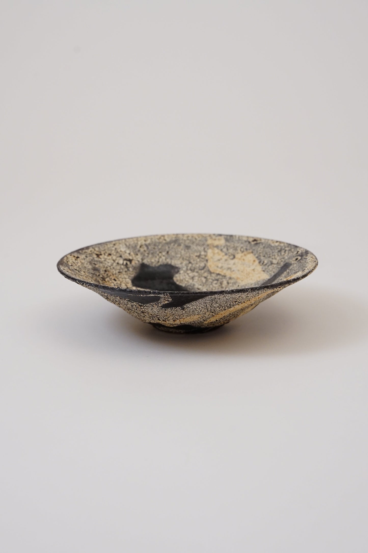 A small handmade ceramic bowl with a textured, speckled glaze in black, gold, and beige, photographed on a neutral light gray background.