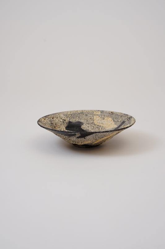 A small handmade ceramic bowl with a textured, speckled glaze in black, gold, and beige, photographed on a neutral light gray background.