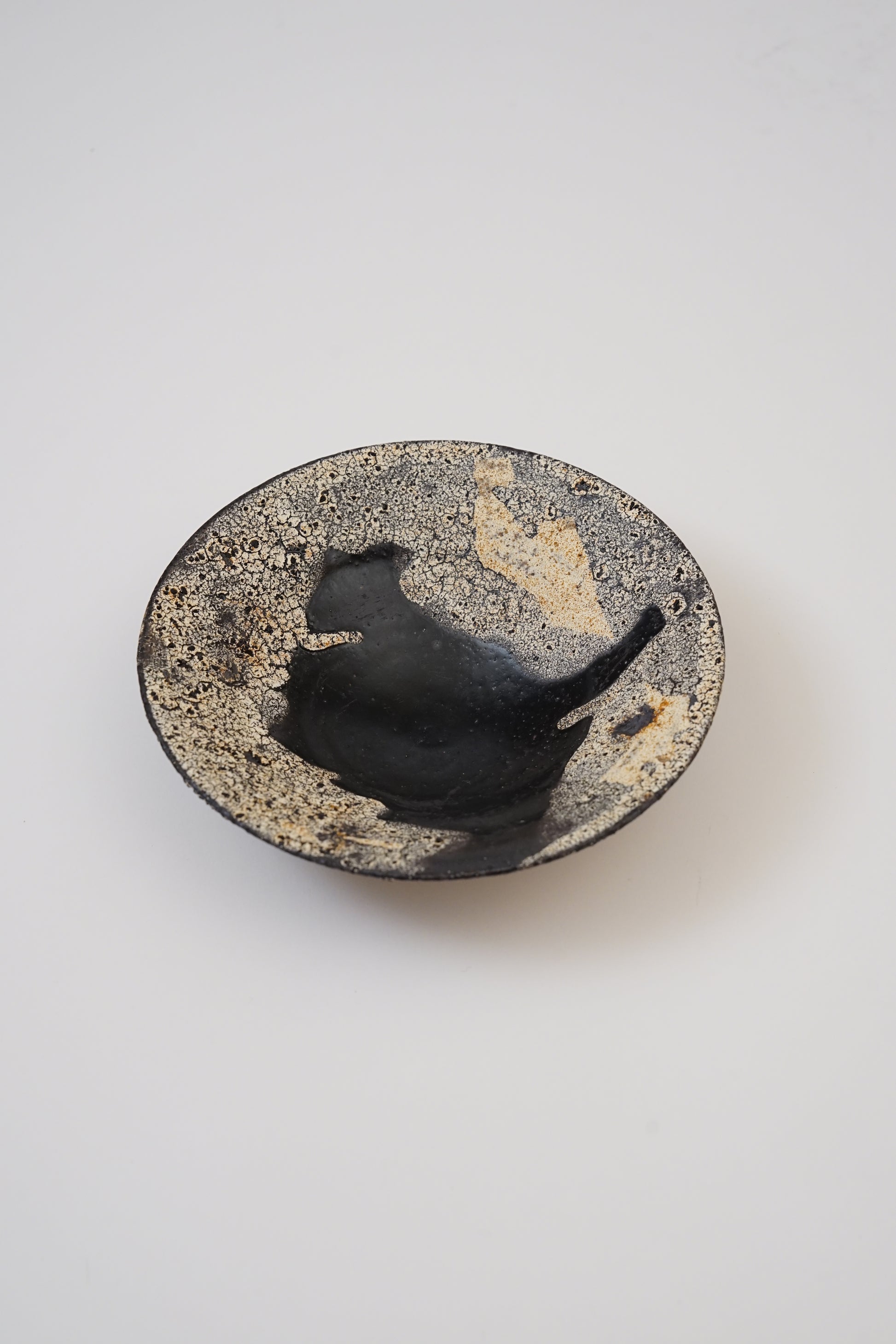 A small handmade ceramic dish with a textured, speckled beige and black exterior and a glossy black interior, photographed from a top-down angle on a light gray background.