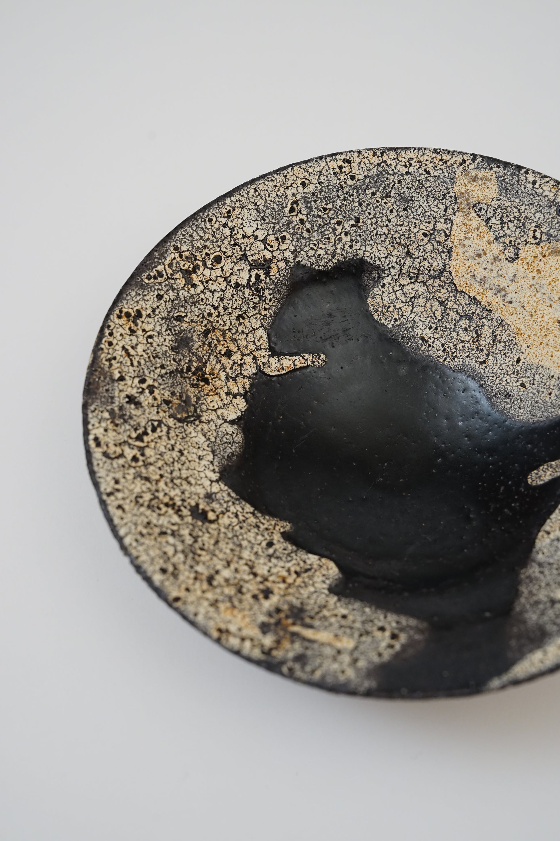 A close-up of a round ceramic bowl or dish with a glossy black center and a textured, speckled beige and brown rim, photographed on a plain light background.