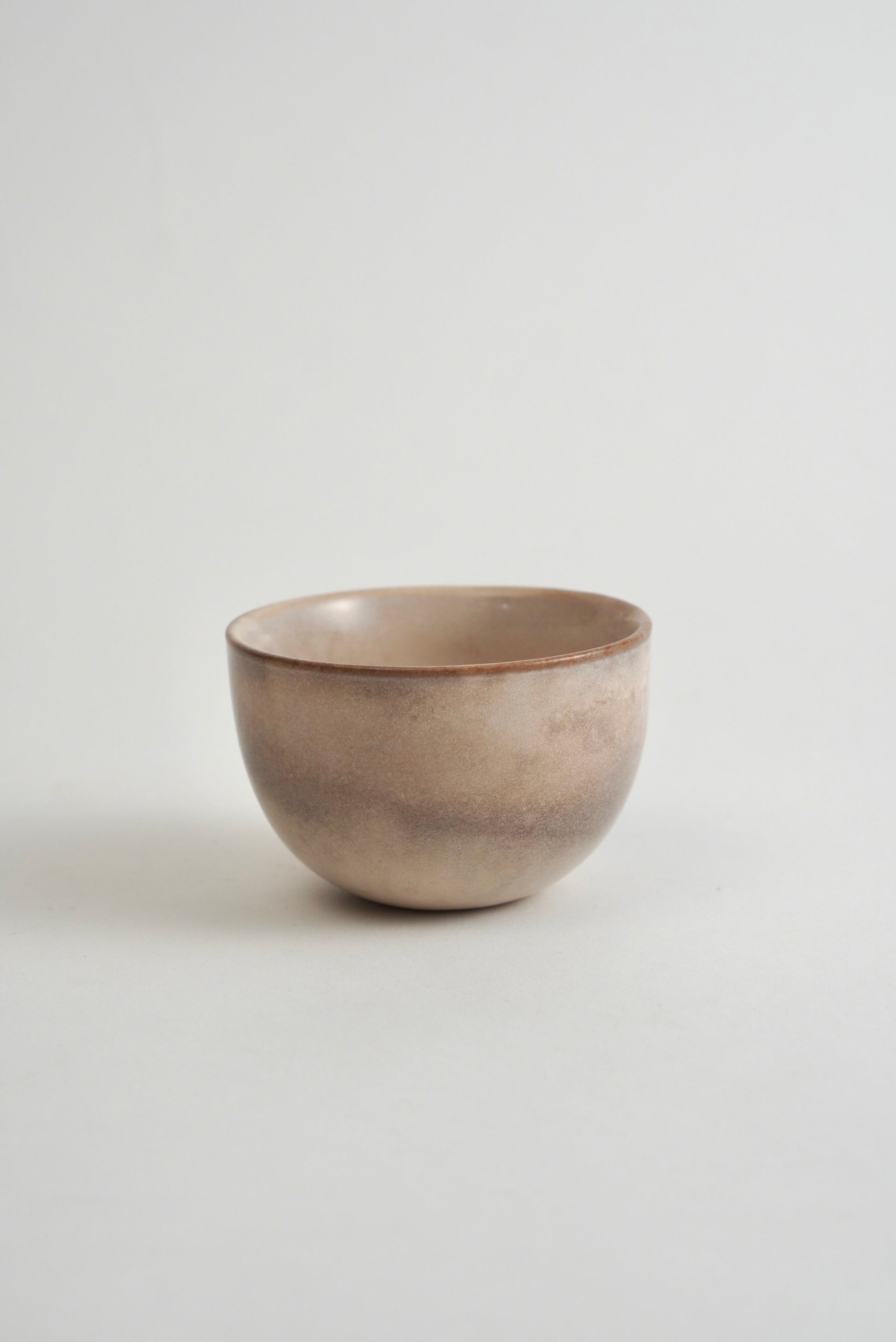 A small handmade ceramic bowl with a textured, earthy finish in shades of beige and brown, set against a plain light gray background.