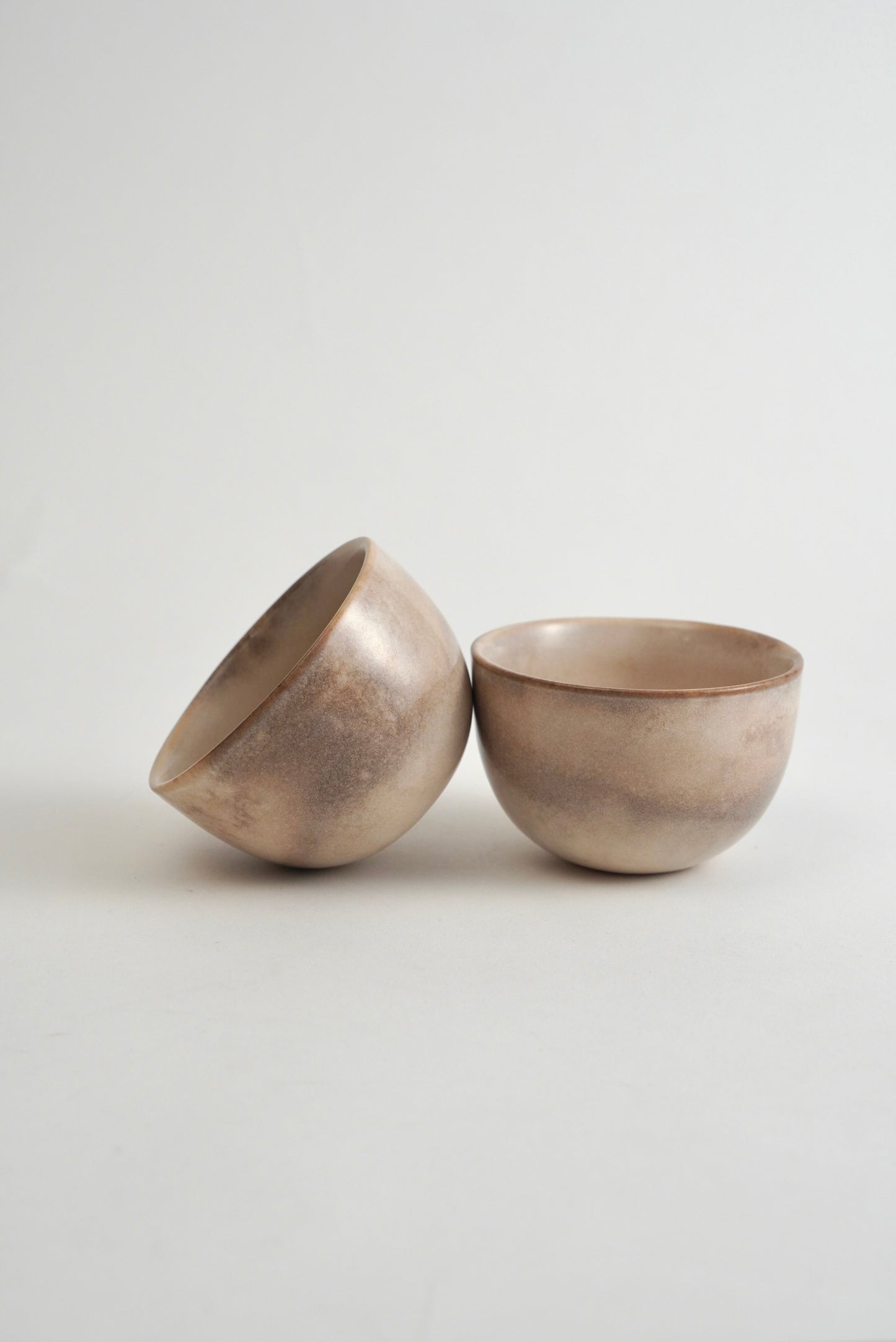 Two small ceramic bowls with a textured, metallic silver finish, one standing upright and the other tilted on its side, set against a plain light gray background.
