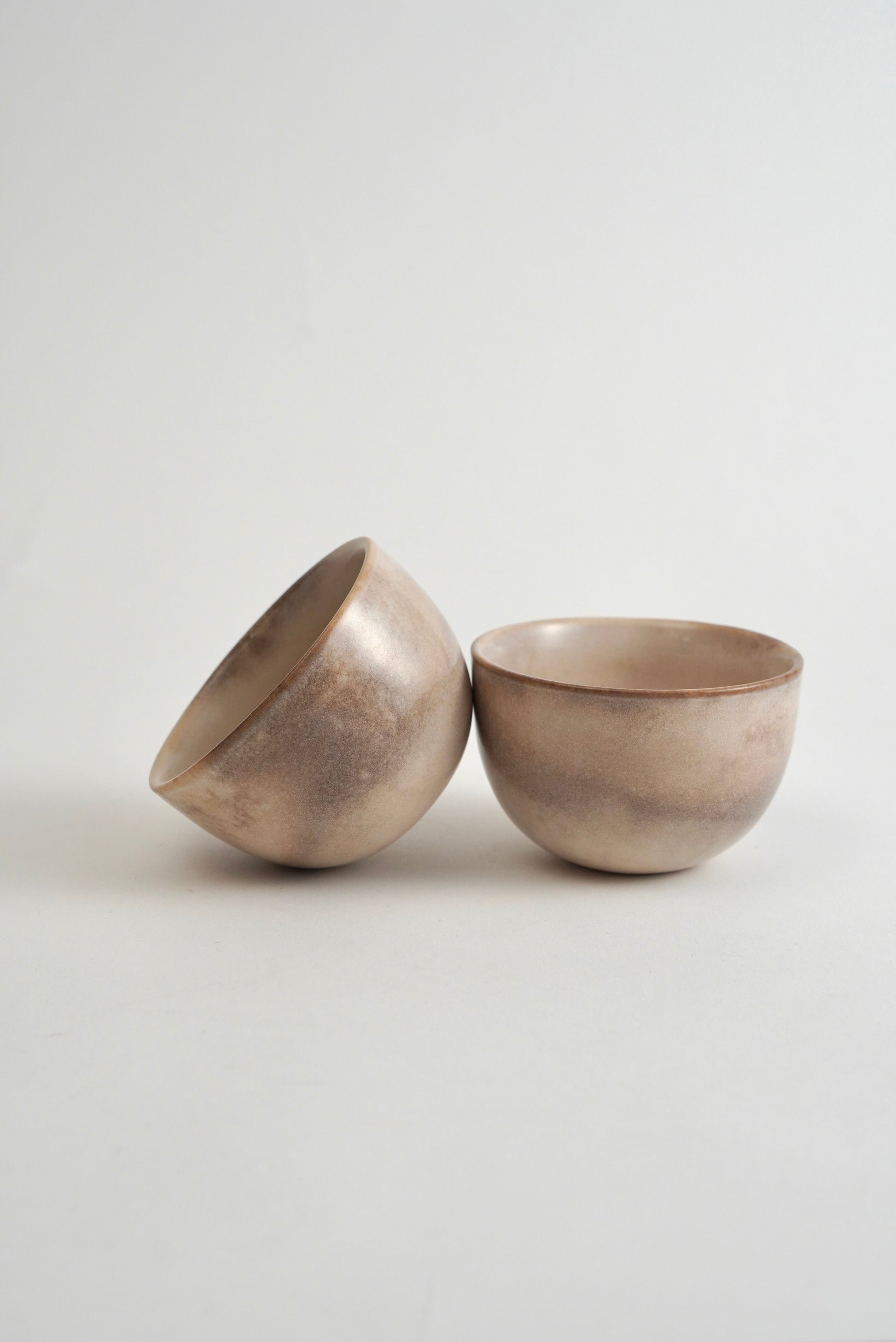 Two small ceramic bowls with a textured, metallic silver finish, one standing upright and the other tilted on its side, set against a plain light gray background.