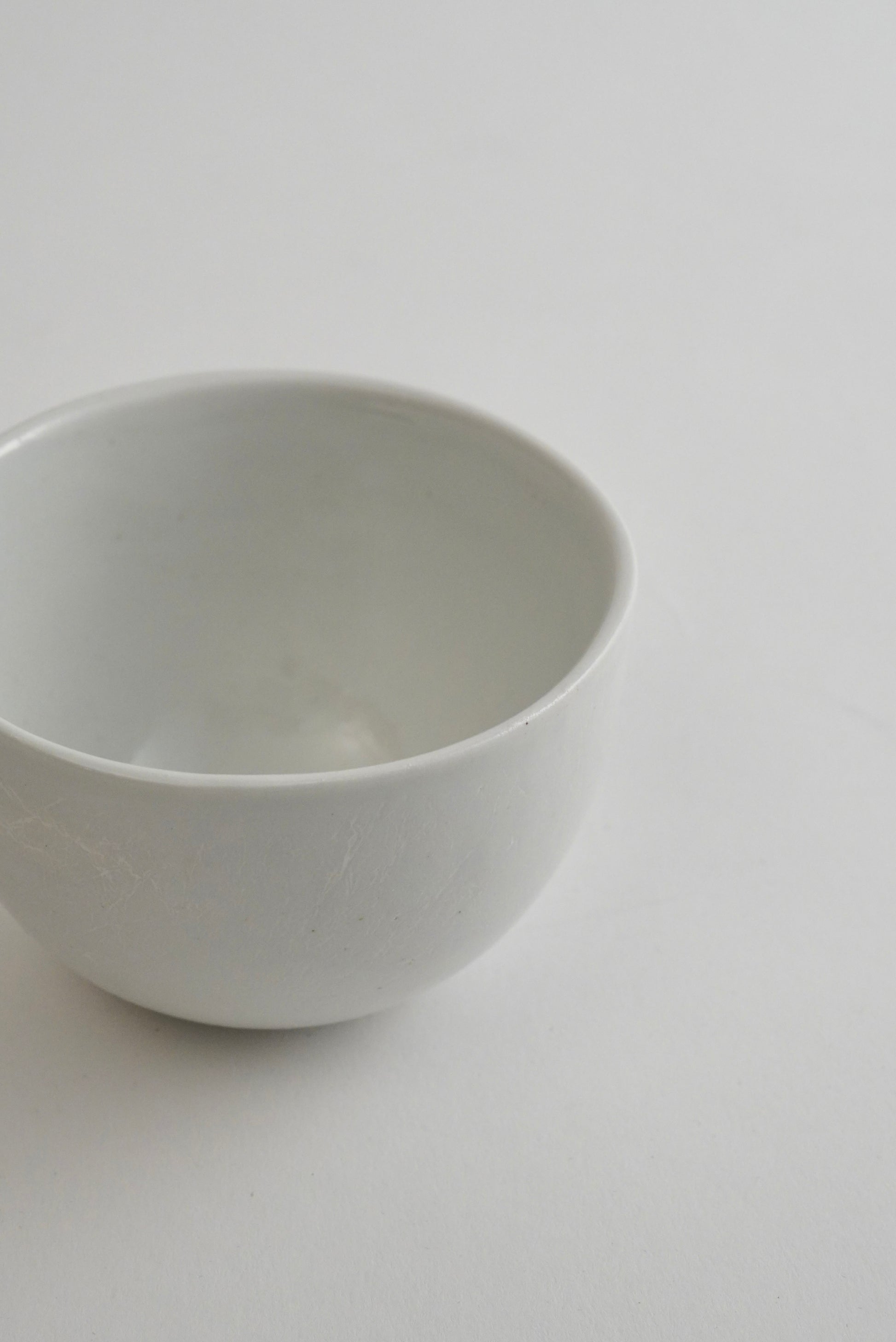 A minimalist white ceramic bowl with a smooth, matte finish, photographed from a high-angle perspective against a clean, light gray background.