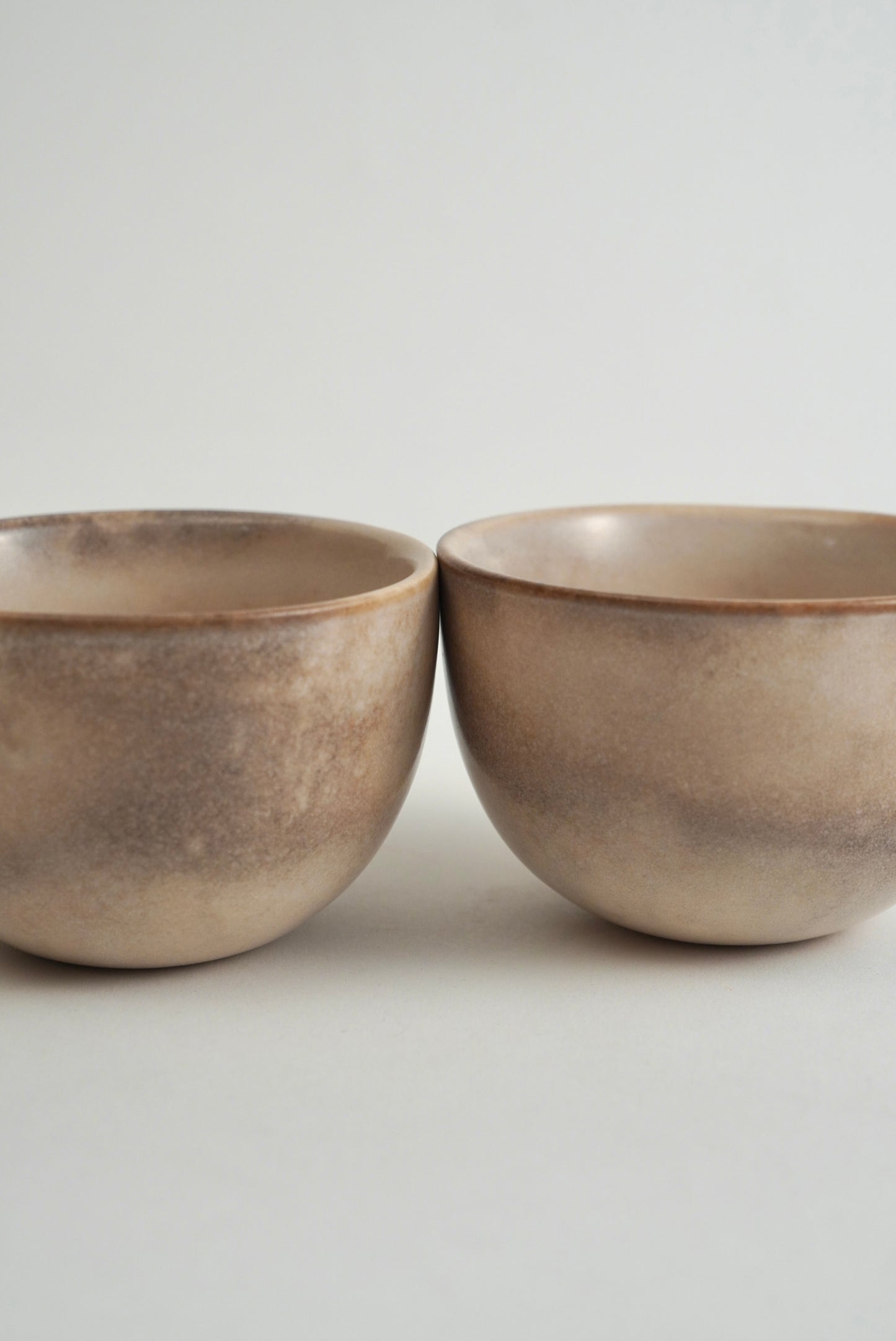 Two small round ceramic bowls with a textured, speckled finish in earthy beige and brown tones, set against a plain light gray background.