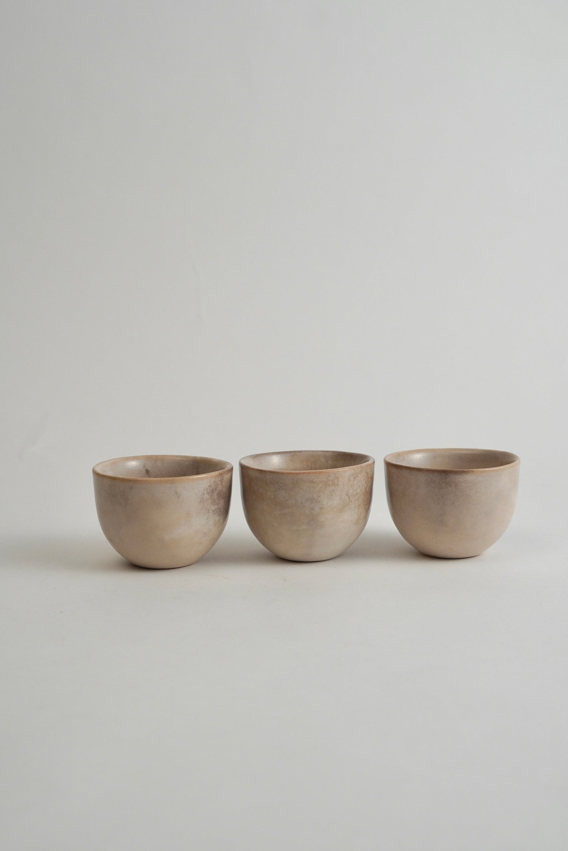 Three small ceramic cups in a matching matte beige finish, arranged side by side on a light gray surface against a neutral gray background.