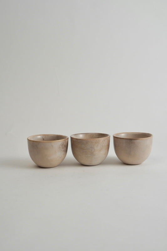 Three small ceramic cups in a matching matte beige finish, arranged side by side on a light gray surface against a neutral gray background.