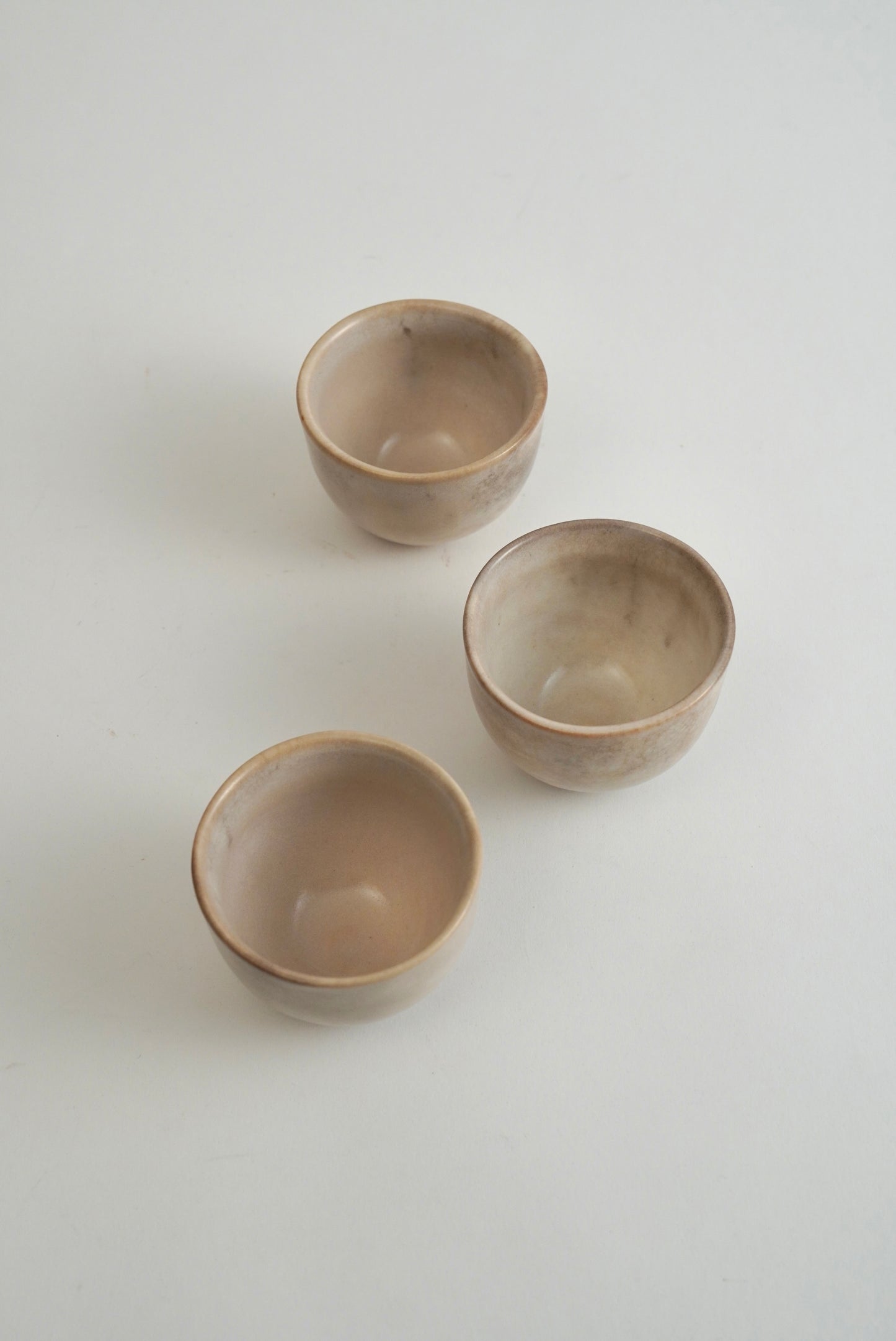 Three small handmade ceramic cups with a matte, speckled beige finish, arranged on a plain light gray background.