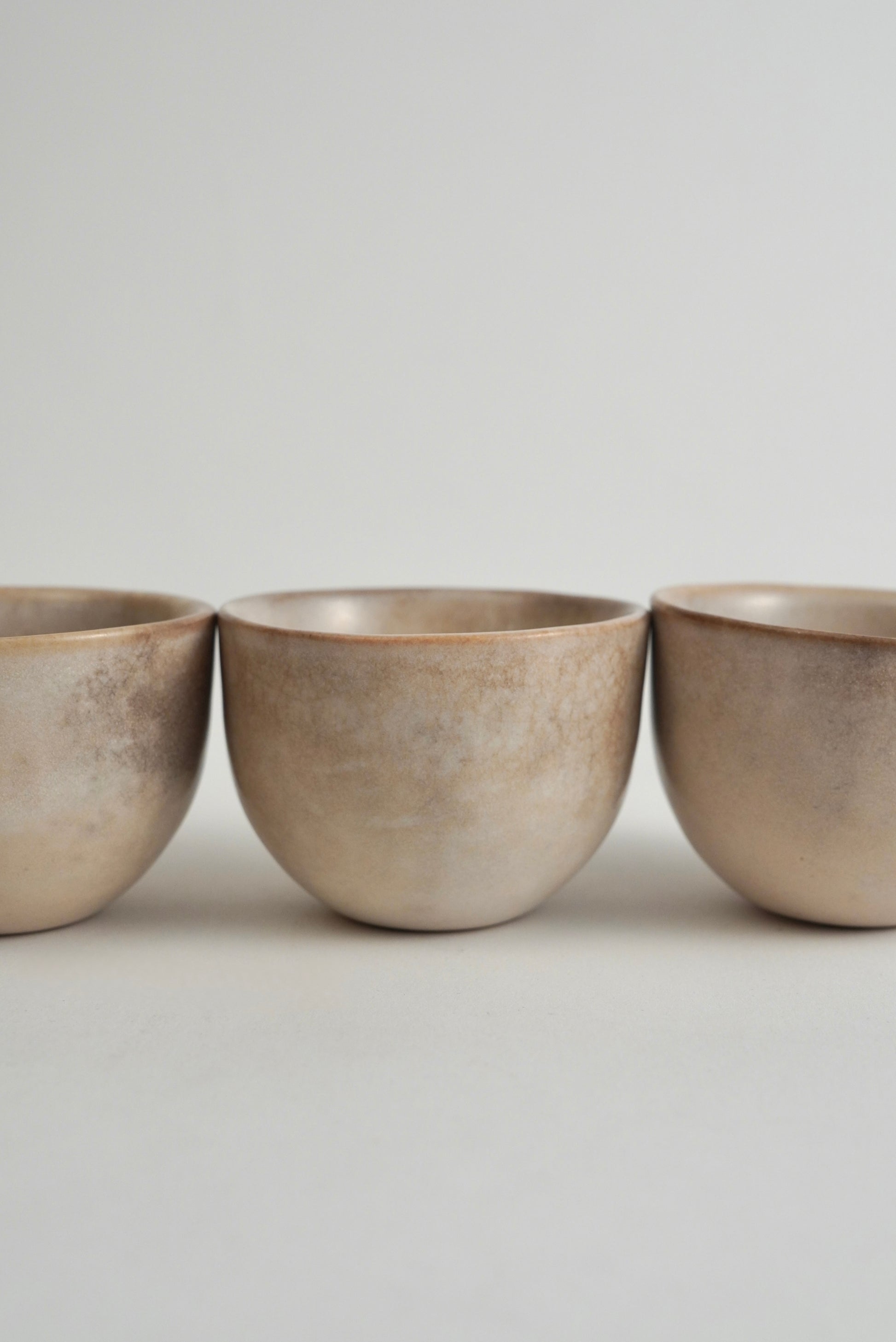 Three small round ceramic cups with a textured, matte metallic finish in a warm beige-bronze tone, arranged in a row against a neutral light gray background.