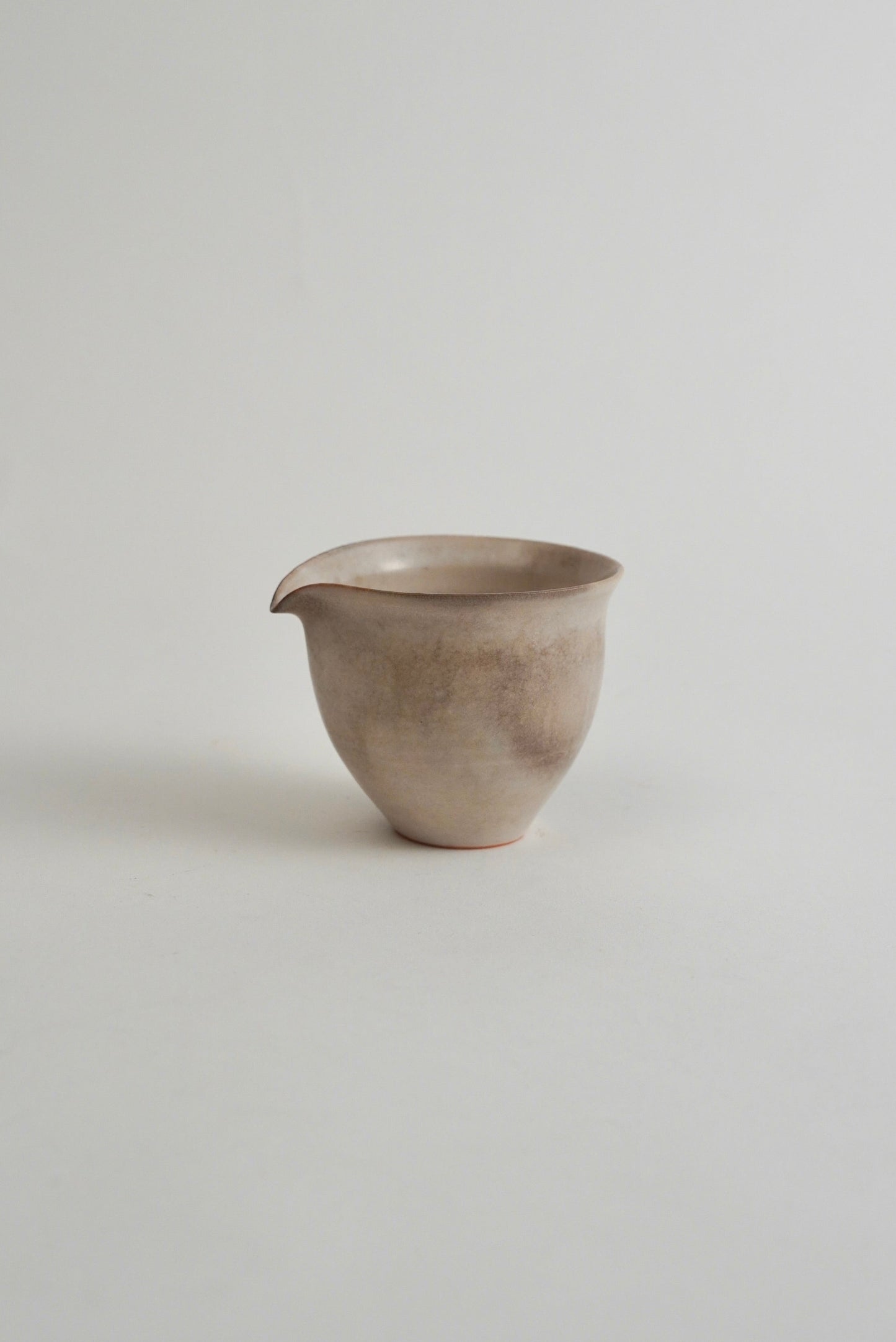 A small handmade ceramic pitcher with a spout, featuring a textured, speckled finish in earthy beige and gray tones, set against a plain light gray background.