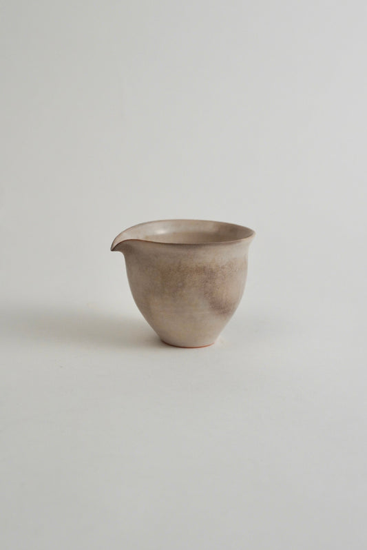 A small handmade ceramic pitcher with a spout, featuring a textured, speckled finish in earthy beige and gray tones, set against a plain light gray background.