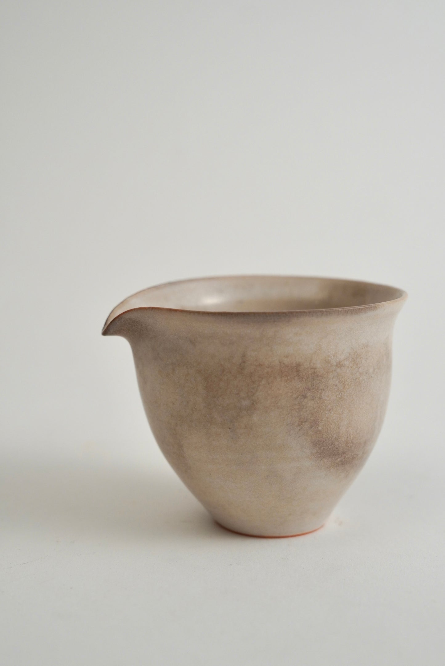 A handmade ceramic pitcher with a small pouring spout, featuring a textured, speckled finish in earthy beige and gray tones, set against a plain light gray background.