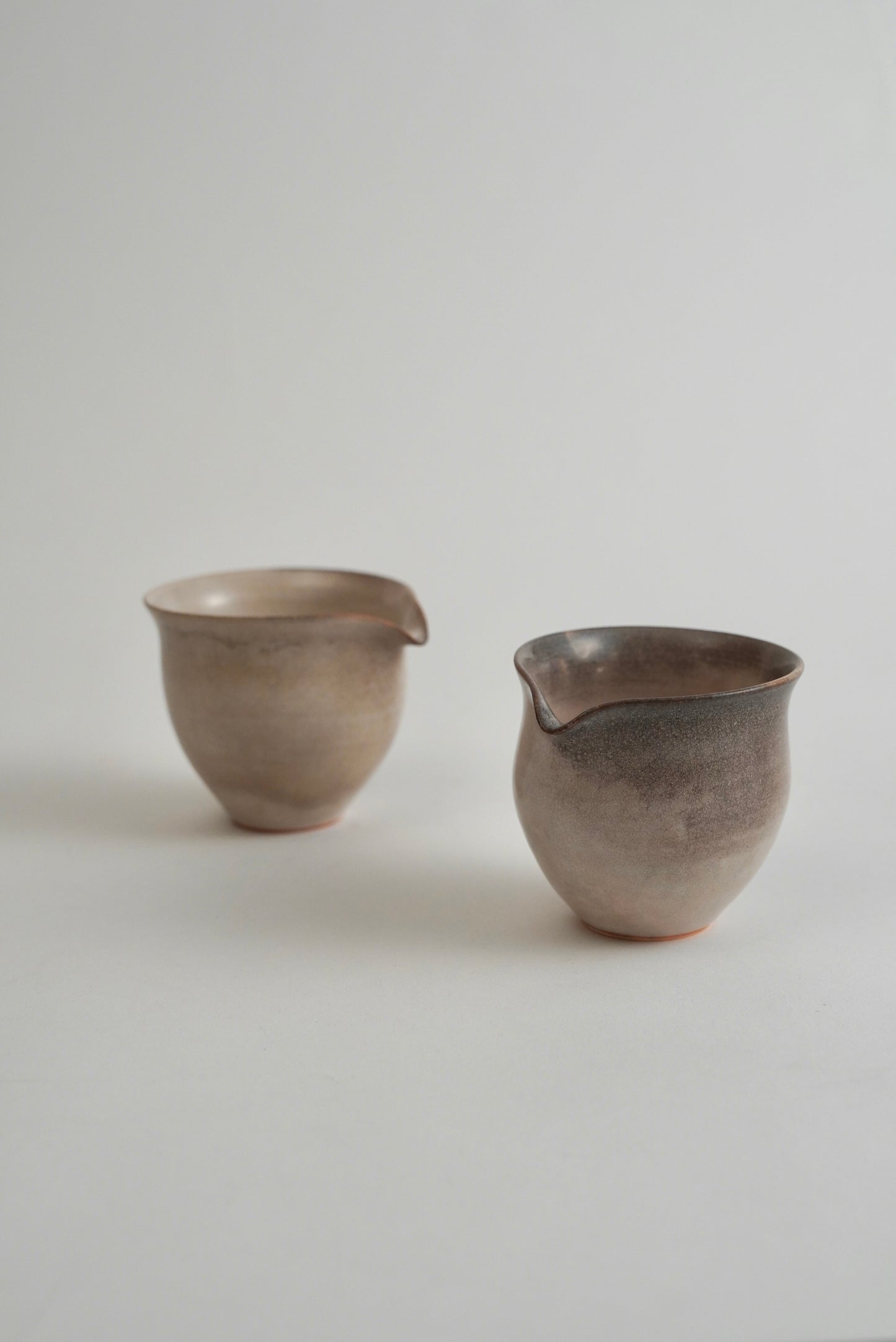 Two handmade ceramic pouring cups with a matte, earthy finish, displayed on a neutral light gray background.